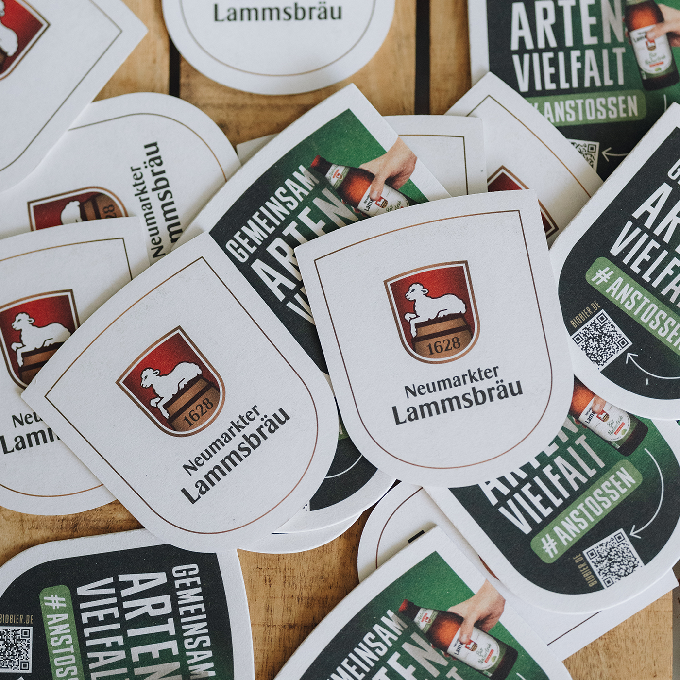 Mehrere wappenförmige Bierdeckel liegen verstreut auf einem Holztisch. Sie zeigen abwechselnd die weiße Seite mit dem Neumarkter Lammsbräu-Logo und eine dunkelgrüne Seite mit dem Slogan „GEMEINSAM ARTEN VIELFALT #ANSTOSSEN“, einem Flaschen-Motiv sowie einem QR-Code.