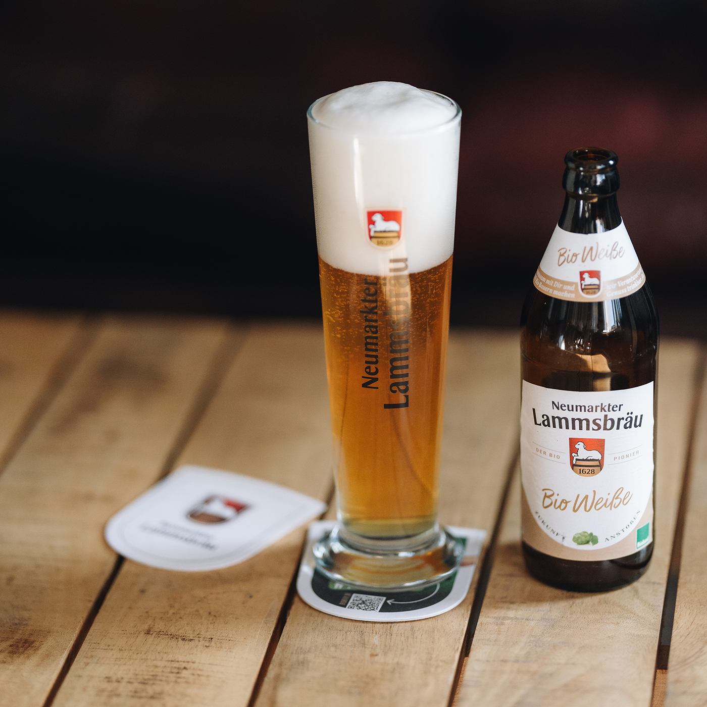 Ein gefülltes Weißbierglas mit üppiger Schaumkrone und vertikalem „Neumarkter Lammsbräu“-Schriftzug steht auf einem wappenförmigen Bierdeckel auf einem Holztisch. Rechts daneben platziert ist eine geöffnete braune Bierflasche der Sorte „Bio Weiße“ mit hellem Etikett.