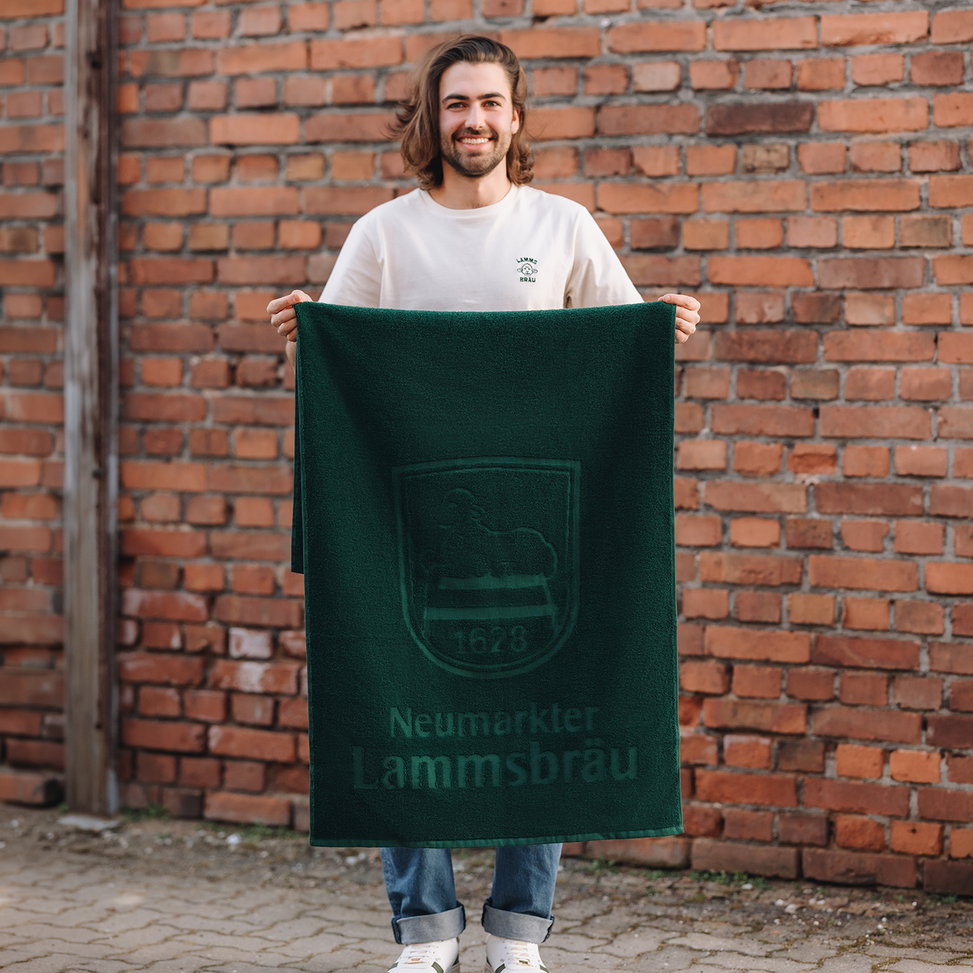 Mann mit braunem Haar und Bart trägt cremefarbenes T-Shirt mit kleinem Lammsbräu-Motiv auf der Brust und hellblaue Jeans vor einer roten Backsteinwand. Er hält ein grünes Baumwoll-Handtuch mit Lammsbräu-Wappen und Schriftzug ausgebreitet vor sich, lächelt in die Kamera.