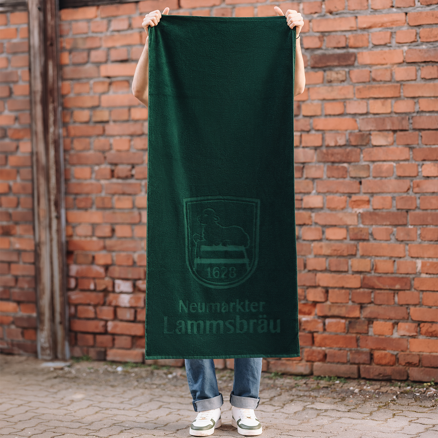 Mann hält ein grünes Baumwoll-Handtuch ausgebreitet vor sich vor einer roten Backsteinwand. Das Handtuch zeigt das Lammsbräu-Logo und den Text "Neumarkter Lammsbräu" in Hoch-Tief-Einwebung. Unter dem Handtuch sichtbar sind hellblaue Jeans und weiße Sneaker mit grünen Akzenten.