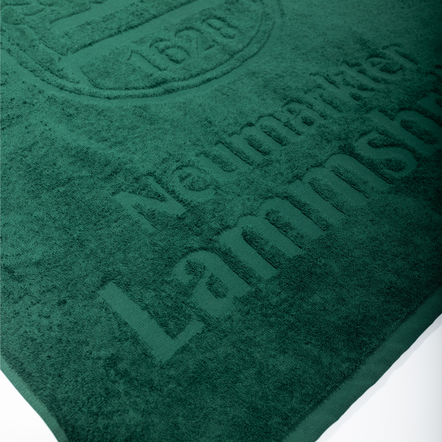 Detailaufnahme eines grünen Baumwoll-Handtuchs. Die Schrift "Neumarkter Lammsbräu" verläuft diagonal über die Oberfläche in dunkelgrüner Farbe. Oben links ist teilweise das Lammsbräu-Wappen sichtbar. Die Handtuch-Textur zeigt die weiche Baumwollstruktur.