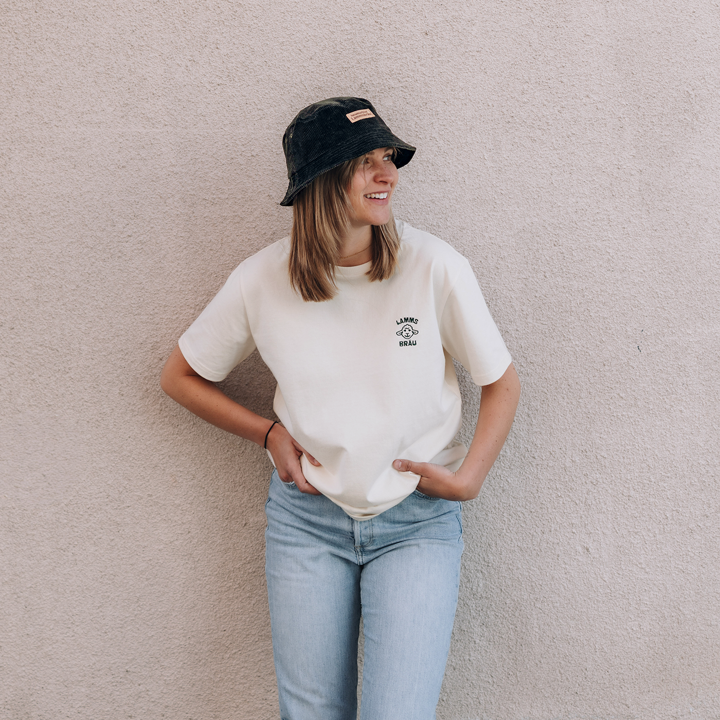 Frau trägt weißes Kurzarm-T-Shirt mit grünem Lammsbräu-Motiv auf der Brust, dazu hellblaue Jeans. Sie trägt eine dunkelgrüne Cord-Bucket-Hat mit einem Lederetikett mit Prägung "Neumarkter Lammsbräu". Die Frau steht vor einer beigen Betonfassade und schaut zur Seite.