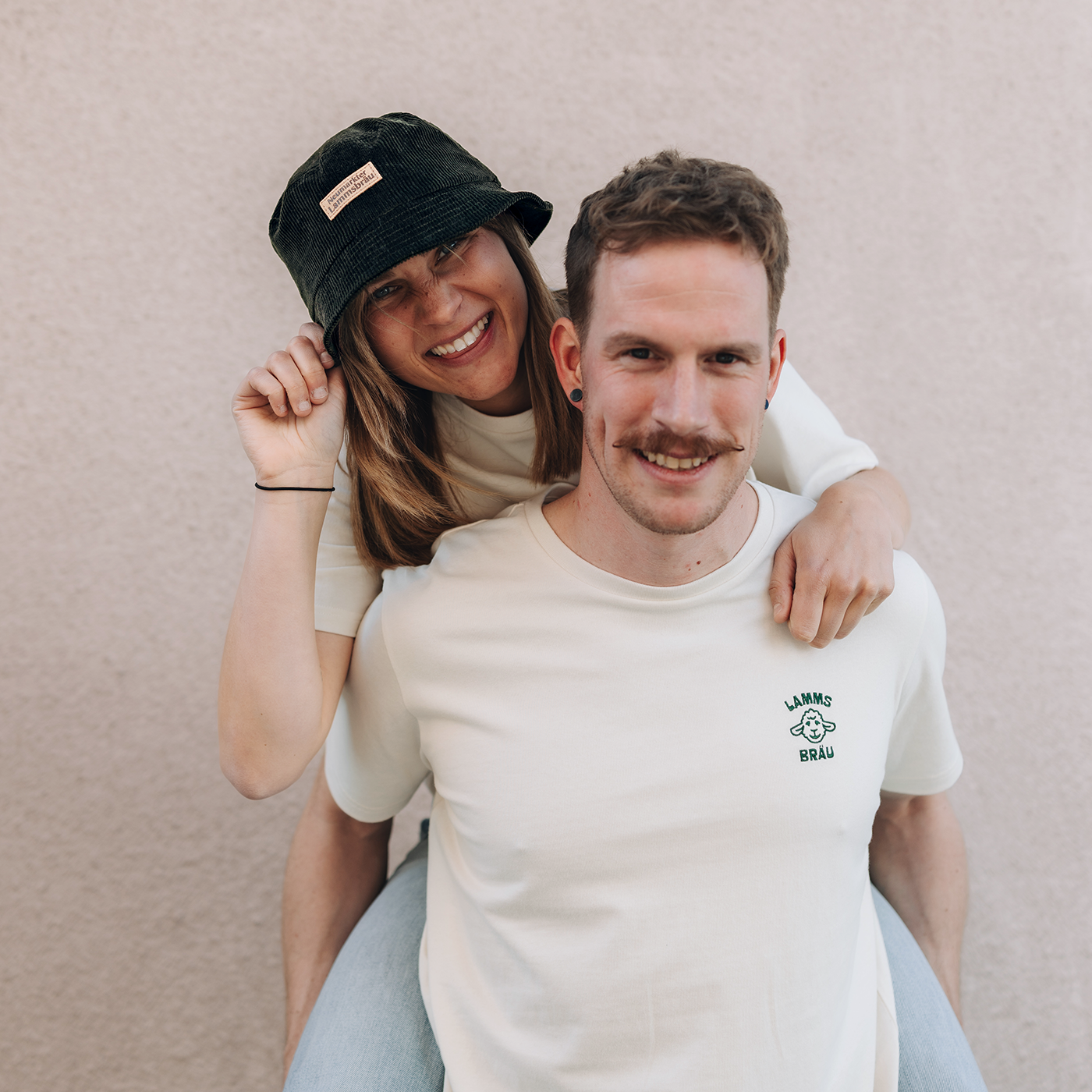 Mann trägt weißes Kurzarm-T-Shirt mit grünem Lammsbräu-Motiv auf der Brust. Eine Frau sitzt auf seinen Schultern, trägt eine dunkelgrüne Cord-Bucket-Hat mit kleinem Lederetikett auf dem der Schriftzug "Neumarkter Lammsbräu" abgebildet ist. Beide lächeln in die Kamera vor neutralem grauem Hintergrund.