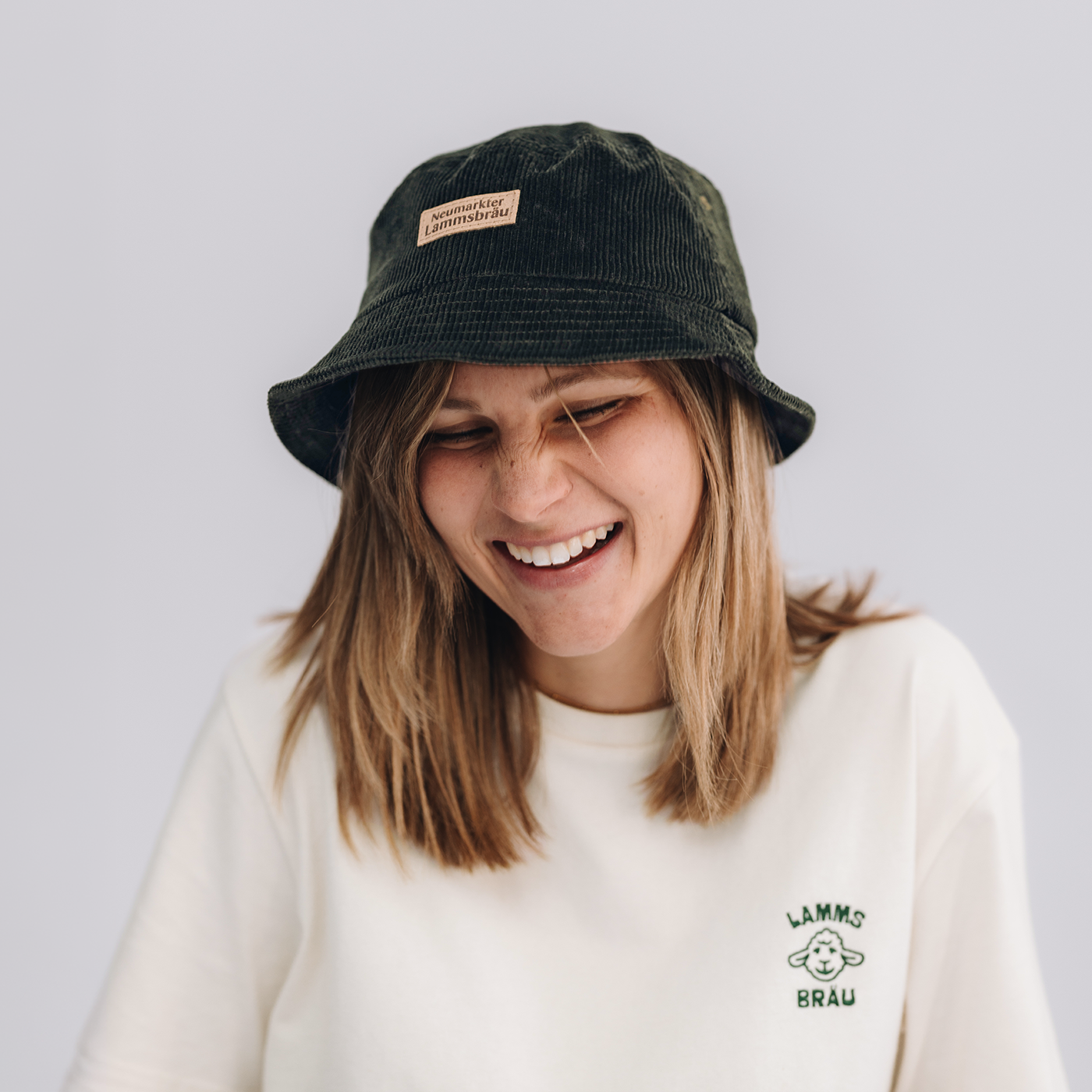 Frau trägt weißes Kurzarm-T-Shirt mit grünem Lammsbräu-Motiv auf der Brust. Sie trägt eine dunkelgrüne Cord-Bucket-Hat mit beigem "Neumarkter Lammsbräu"-Label, schaut nach unten und lächelt vor neutralem grauem Hintergrund.