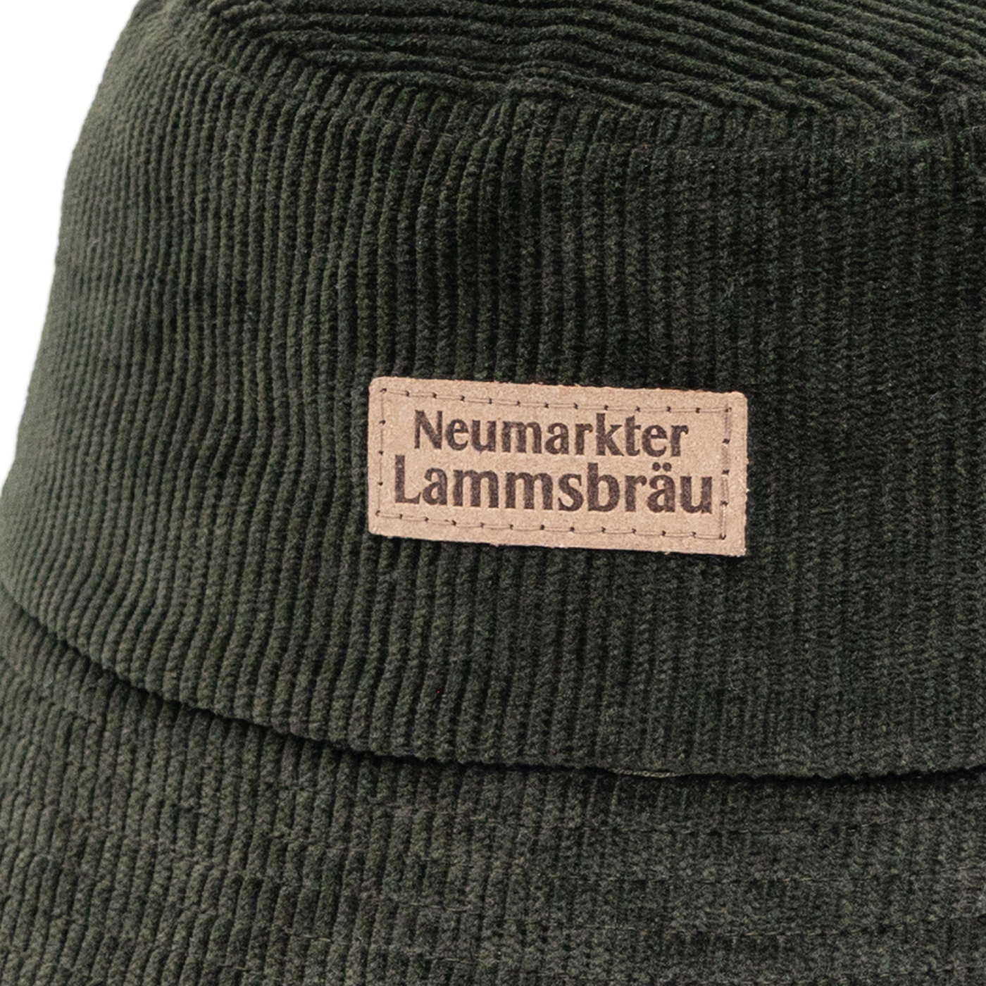 Detailaufnahme einer grünen Cord-Bucket-Hat mit beigefarbenem rechteckigem Leder-Label. Das Label trägt die schwarze Aufschrift "Neumarkter Lammsbräu" und ist mit dekorativen Stichlinien verziert. Darunter die gerippte grüne Stofftextur des Hutes.