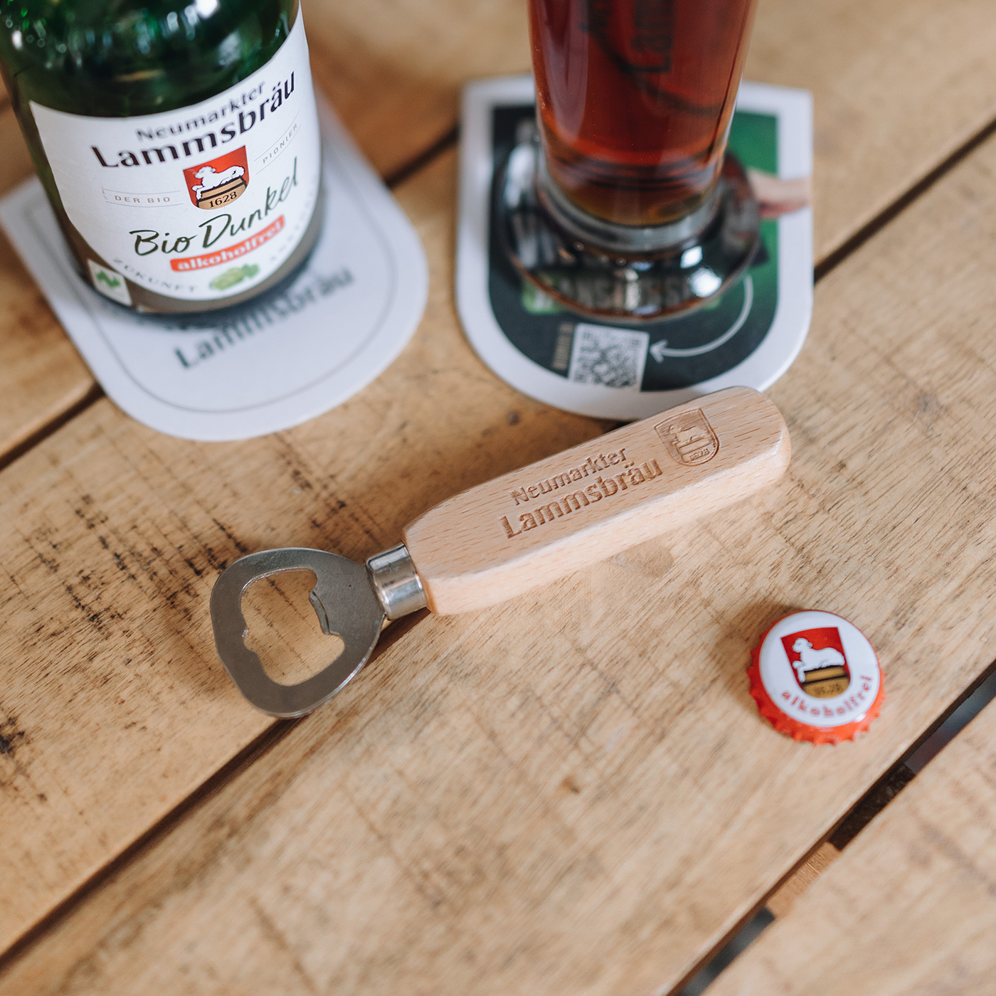 Flach liegende Arrangierung auf hellem Holztisch mit Neumarkter Lammsbräu-Produkten. Links oben eine dunkelbraune Bierflasche mit beigefarbenem Label „Bio Dunkel alkoholfrei", rechts oben ein gefülltes Glas mit dunkelrotem Bier auf weiß-grünem Bierdeckel. In der Bildmitte ein Holz-Flaschenöffner mit eingraviertem „Neumarkter Lammsbräu"-Schriftzug und metallischer Öffnungsfunktion. Rechts unten ein roter Kronkorken mit Lammsbräu-Logo.