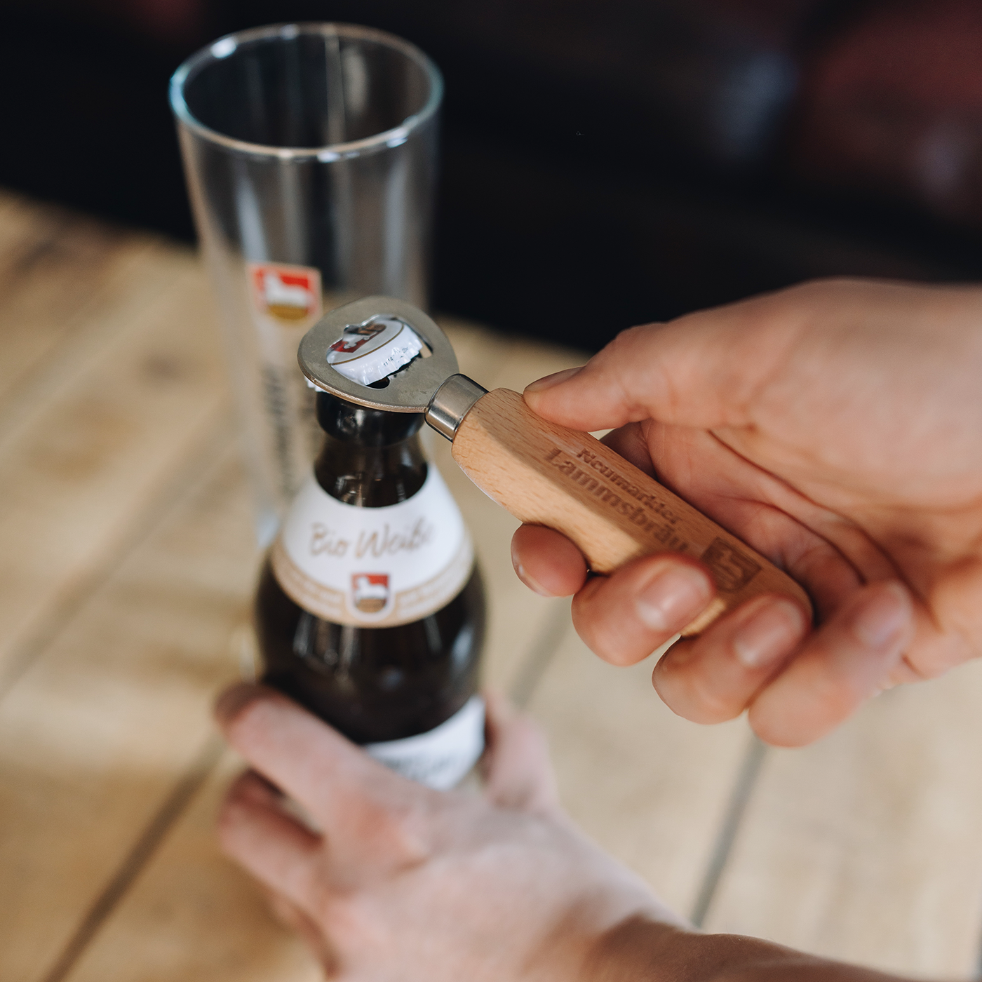 Nahaufnahme einer Hand, die einen Holz-Flaschenöffner mit eingraviertem „Neumarkter Lammsbräu"-Schriftzug und Wappen hält und damit eine dunkelbraune Bierflasche mit weißem Etikett öffnet. Im Hintergrund unscharfes leeres Glas mit rotem Wappen-Logo auf hellem Holztisch.