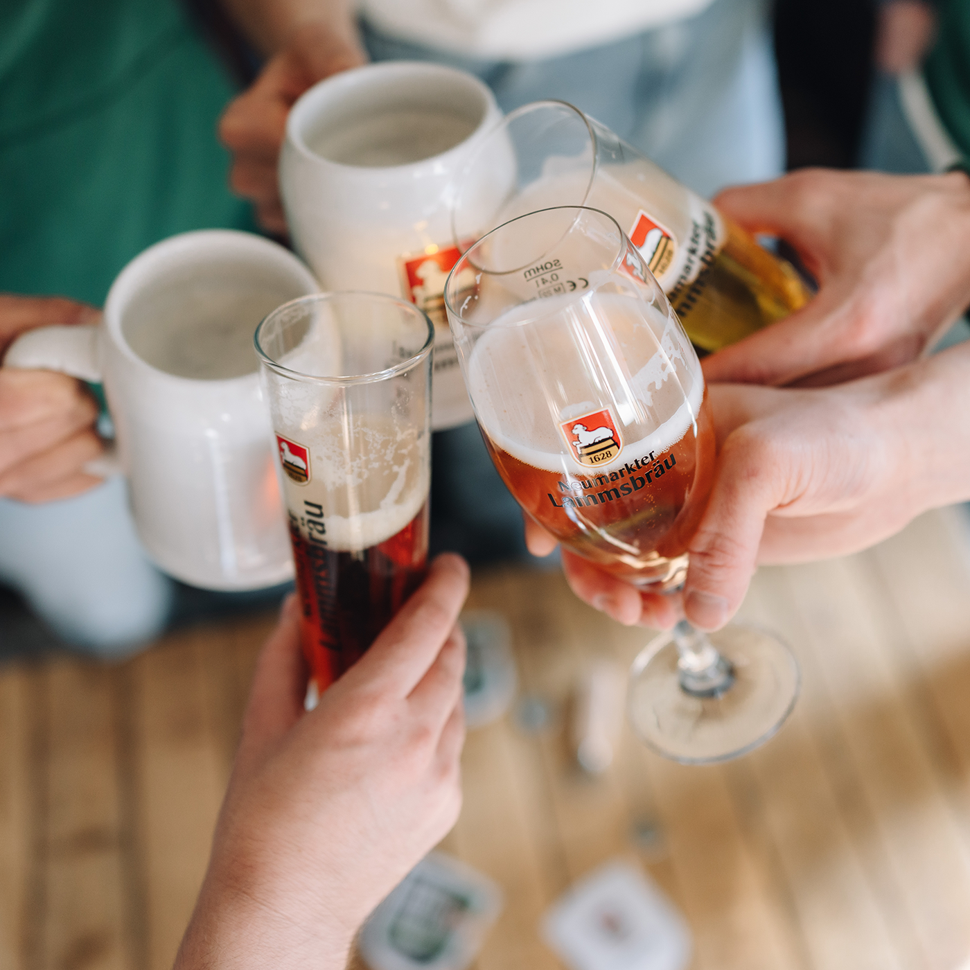 Nahaufnahme von fünf Händen, die verschiedene Gläser mit Lammsbräu-Schriftzug halten. Drei Gläser zeigen das Lammsbräu-Wappen. Zwei weiße Keramikkrüge sind im Hintergrund verschwommen sichtbar. Im Vordergrund ein Holztisch mit unscharfen Bierdeckeln.