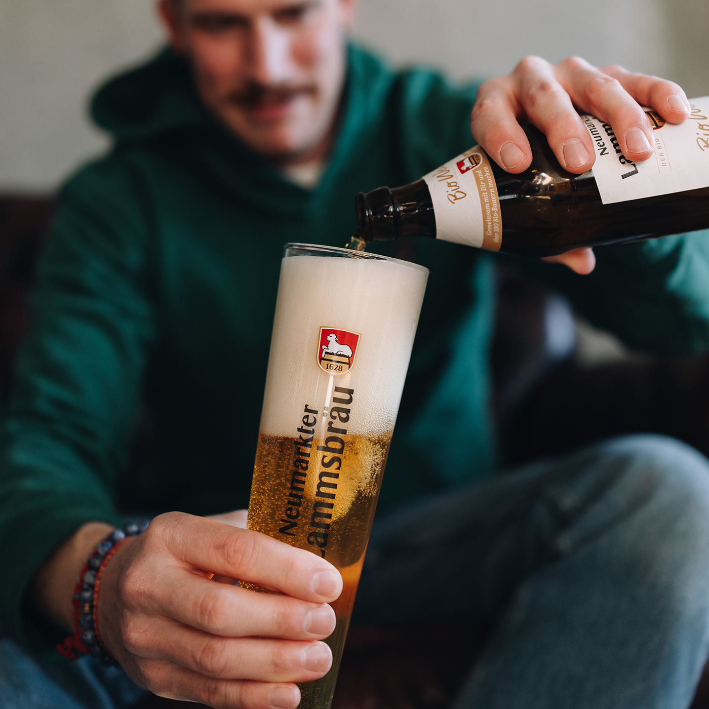Mann in grünem Kapuzen-Sweatshirt mit kleinem Lammsbräu-Logo gießt Bier aus einer dunklen Lammsbräu-Flasche in ein Glas mit "Neumarkter Lammsbräu"-Schriftzug. Das Glas ist halb gefüllt mit hellem schäumendem Bier. Der Mann blickt auf das einschenken herab.