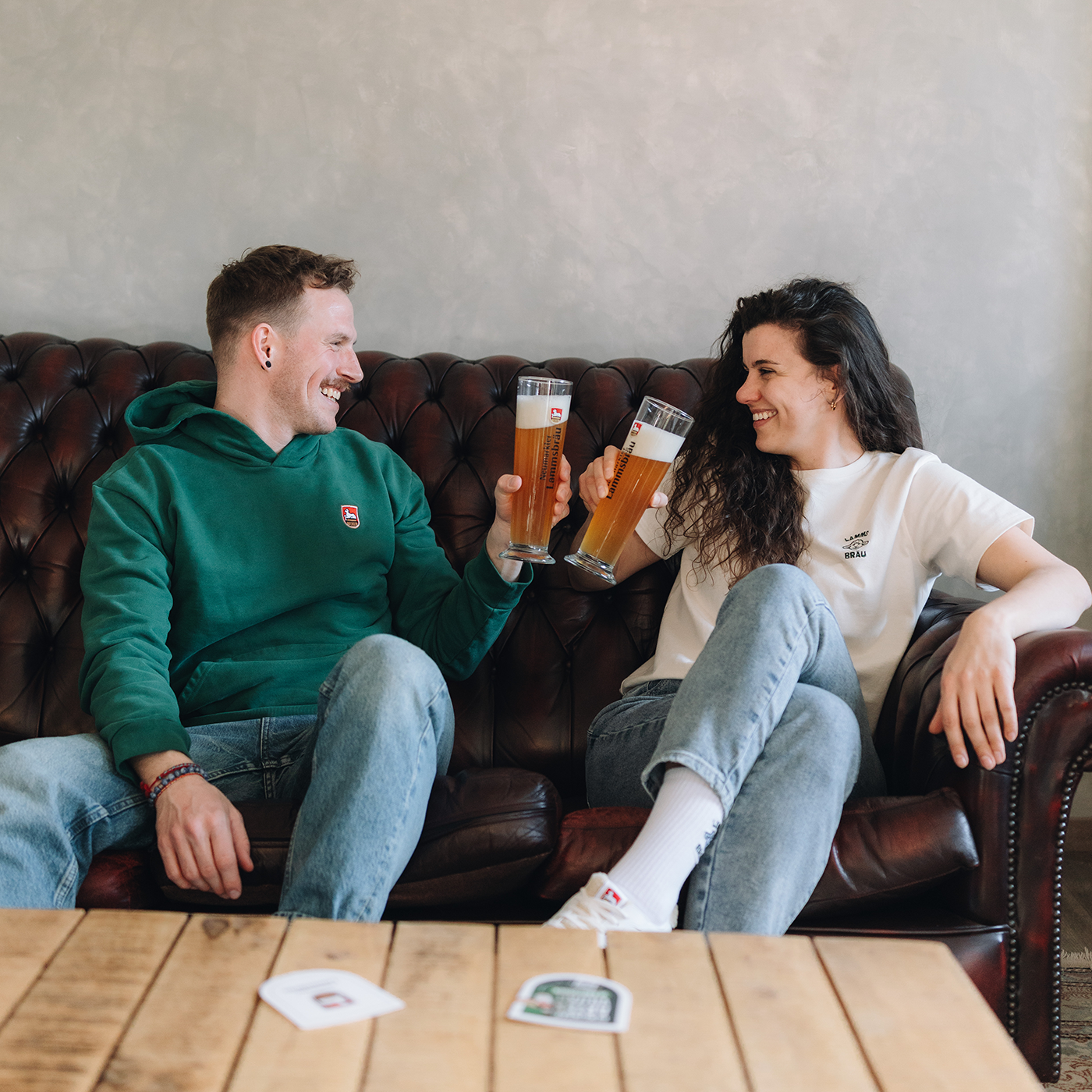 Mann in grünem Kapuzen-Sweatshirt und Frau in weißem T-Shirt mit Lammsbräu-Motiv sitzen nebeneinander auf einem dunkelbraunen Ledersofa. Sie halten jeweils ein Bierglas mit "Neumarkter Lammsbräu"-Aufdruck gefüllt mit Bier, stoßen an und lächeln sich gegenseitig an. Im Hintergrund weiße Wand und Holztisch.