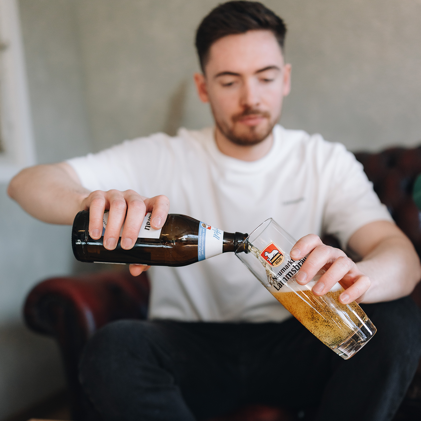 Mann in weißem T-Shirt sitzt auf dunklem Ledersofa und gießt aus einer dunkelbraunen Lammsbräu-Bierflasche mit blauem Label in ein Glas mit Lammsbräu-Wappen und Schriftzug. Das Glas ist halb gefüllt mit schäumendem Bier. Der Mann blickt auf das Einschenken herab. Im unscharfen Hintergrund das braune Sofa und helle Wand.