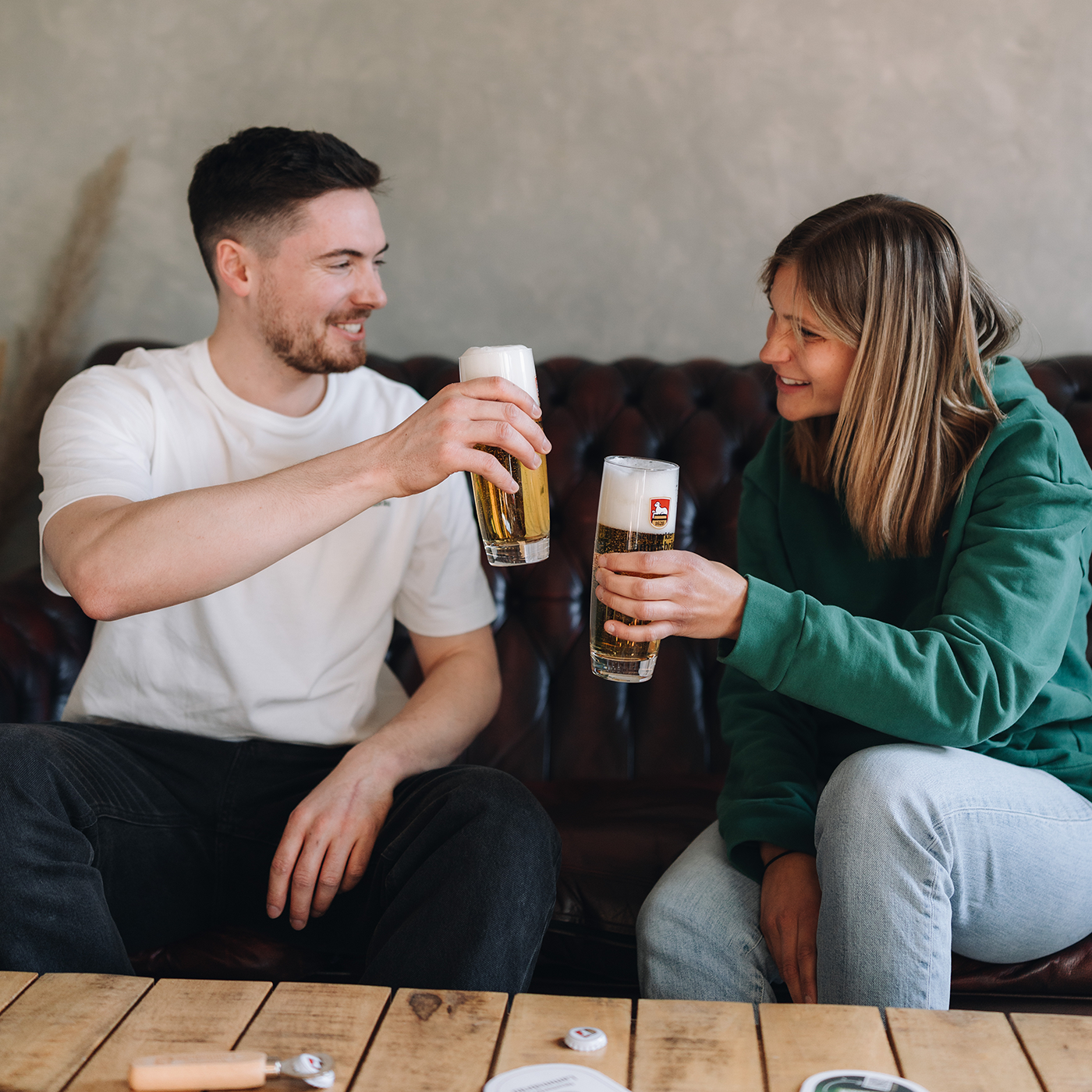 Mann in weißem T-Shirt und Frau in grünem Kapuzen-Sweatshirt sitzen auf einem dunkelbraunen Ledersofa und stoßen mit zwei gefüllten Biergläsern an. Beide Gläser enthalten Lammsbräu-Bier mit weißem Schaum. Die beiden blicken sich lächelnd an. Im Vordergrund sichtbar ein Holztisch mit zwei Bierdeckeln. Im Hintergrund eine graue Wand.