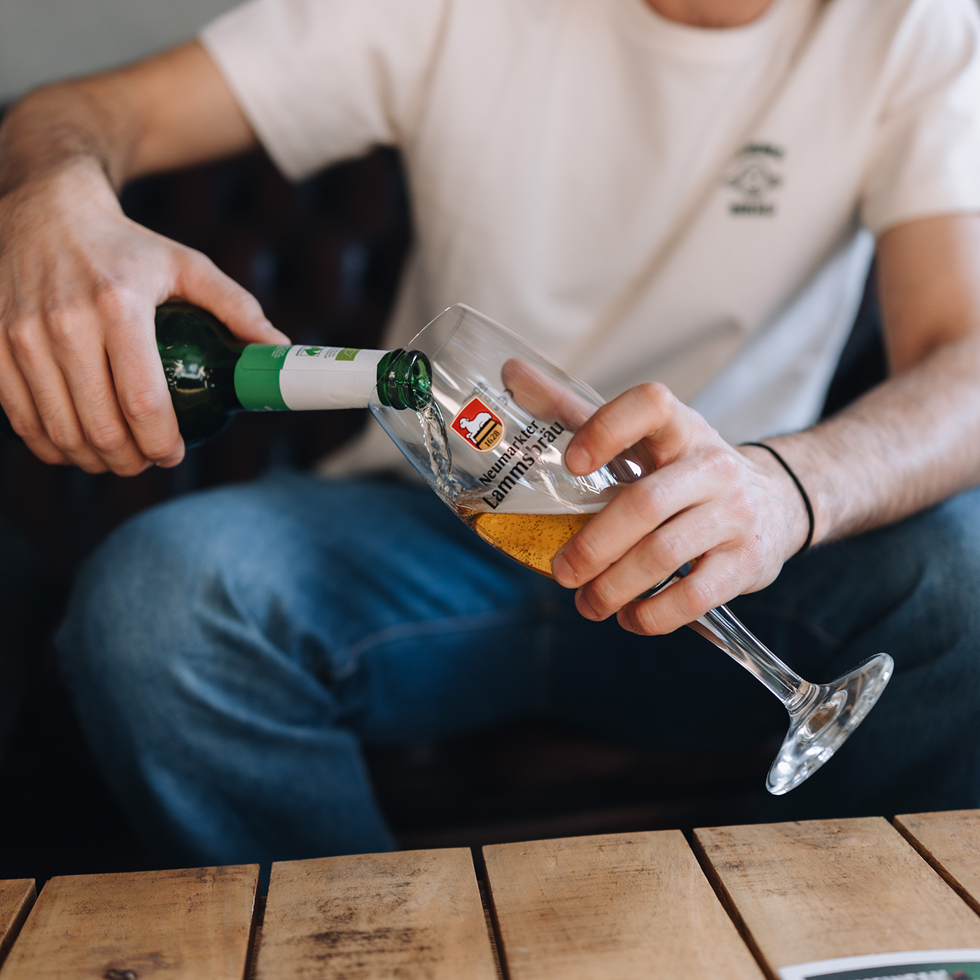 Nahaufnahme von zwei Händen einer Person, die eine grüne Lammsbrau-Bierflasche mit weißem Label in ein zylindrisches Glas mit rotem Lammsbräu-Wappen und schwarzem Schriftzug gießt. Das Glas ist halb gefüllt mit goldenem Bier. Die Person trägt ein cremefarbenes T-Shirt mit kleinem Lammsbräu-Motiv und blaue Jeans. Im Hintergrund unscharfer dunkler Sessel und Holztisch.