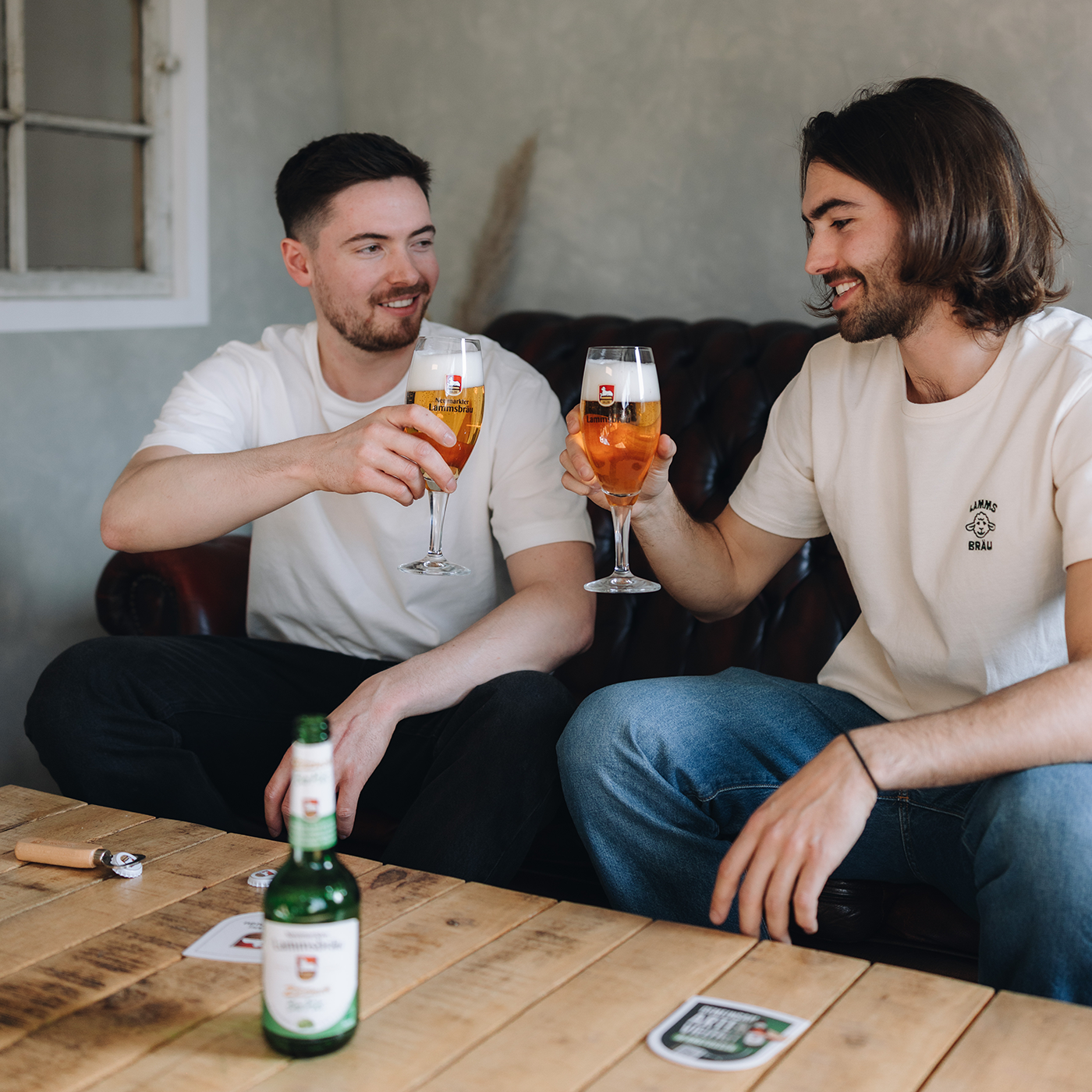 Zwei Männer sitzen auf einem dunkelbraunen Ledersofa vor einer hellen Wand mit weißem Fenster. Der linke Mann trägt ein weißes T-Shirt, der rechte Mann ein cremefarbenes T-Shirt mit Lammsbräu-Motiv. Beide halten elegante Gläser mit goldenem Lammsbräu-Bier und weißem Schaum und blicken sich lächelnd an. Auf dem Holztisch davor steht eine grüne Lammsbräu-Bierflasche mit weißem Label, daneben liegen Bierdeckel.