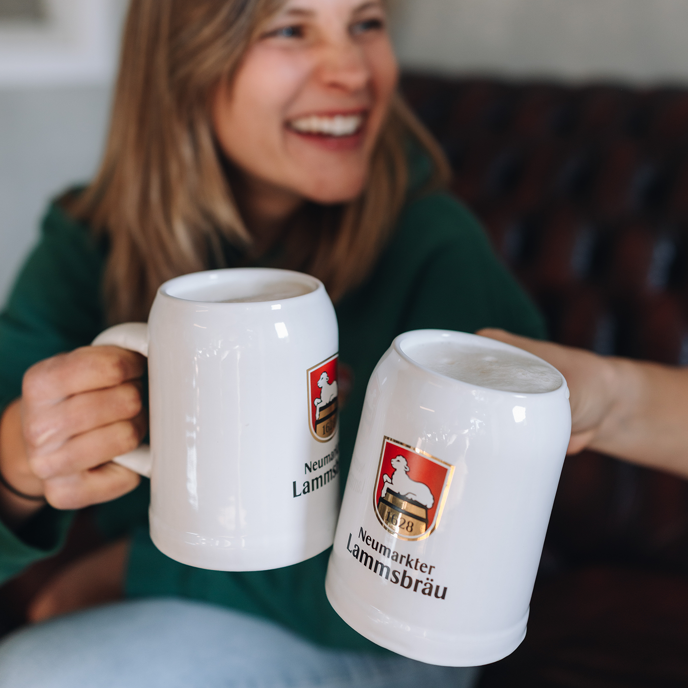 Nahaufnahme zweier anstoßender weißer Keramikkrüge, auf denen das Neumarkter Lammsbräu-Logo mit Wappen und Schriftzug abgedruckt ist. Im unscharfen Hintergrund blickt eine lächelnde Frau in einem grünen Kapuzenpullover auf die Krüge.