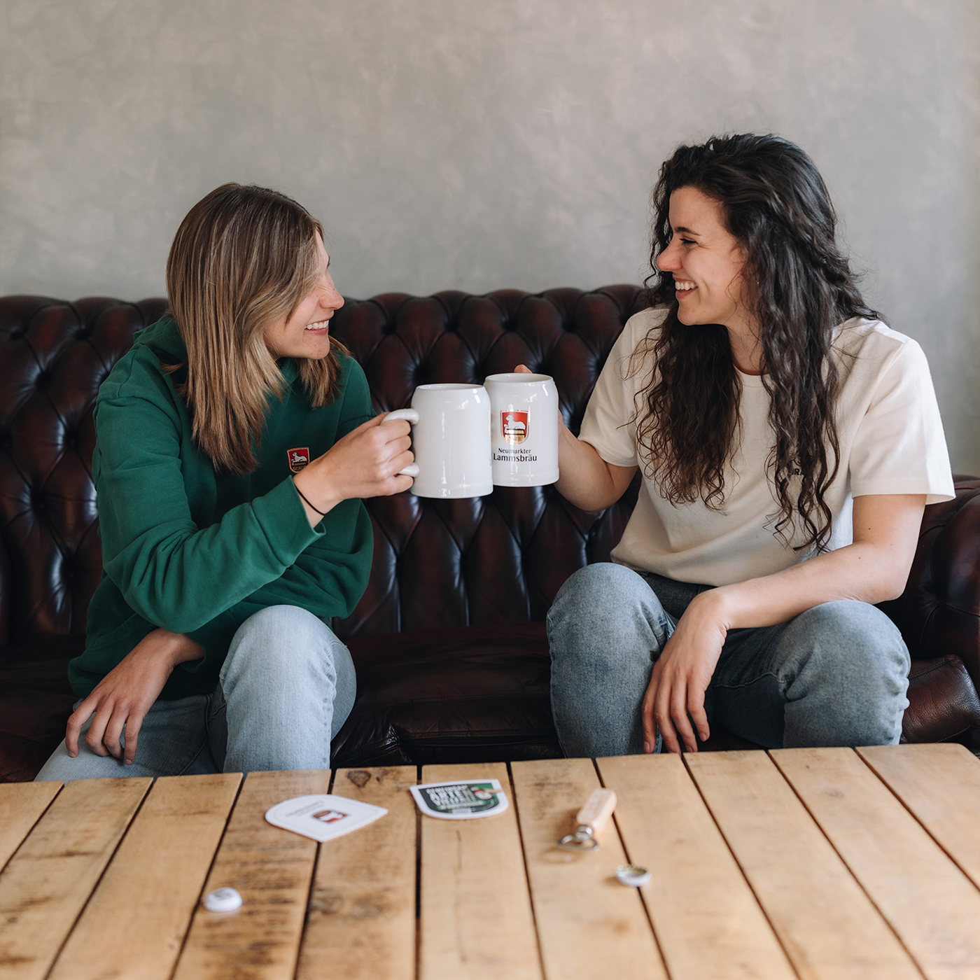 Zwei Frauen sitzen auf einem dunklen Ledersofa und stoßen lächelnd mit zwei weißen Keramikkrügen an, die das Neumarkter Lammsbräu-Logo tragen. Die linke Frau trägt einen grünen Kapuzenpullover mit kleinem Logo-Patch, im Vordergrund befindet sich ein Holztisch mit Bierdeckeln und Flaschenöffner.