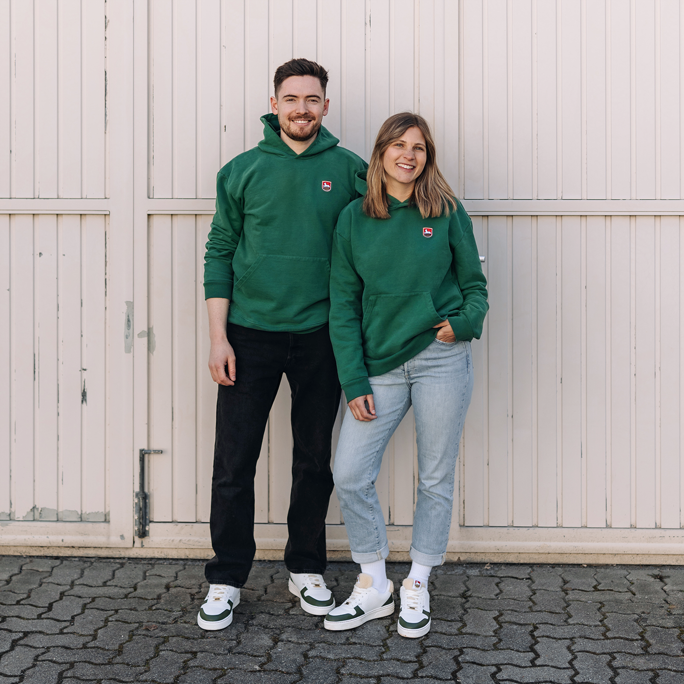 Mann und Frau stehen nebeneinander vor einer weißen Wellblechfassade mit vertikalen Rillen. Beide tragen grüne Kapuzen-Sweatshirts mit kleinem Lammsbräu-Wappen auf der Brust. Der Mann trägt eine schwarze Hose, die Frau hellblaue Jeans. Beide lächeln in die Kamera.