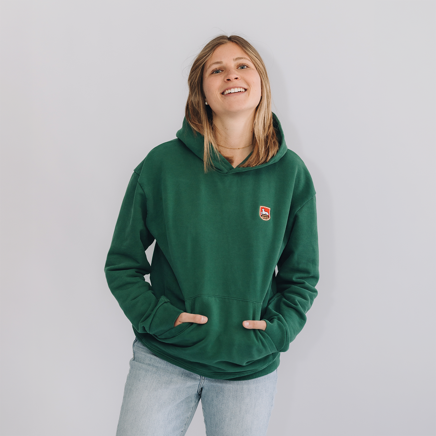 Frau trägt grünes Kapuzen-Sweatshirt mit einem kleinen gestickten Lammsbräu-Wappen auf der Brust und hellblaue Jeans, Vorderansicht auf neutralem grauem Hintergrund. Ihre Hände befinden sich in den Hosentaschen, sie schaut nach oben und lächelt.