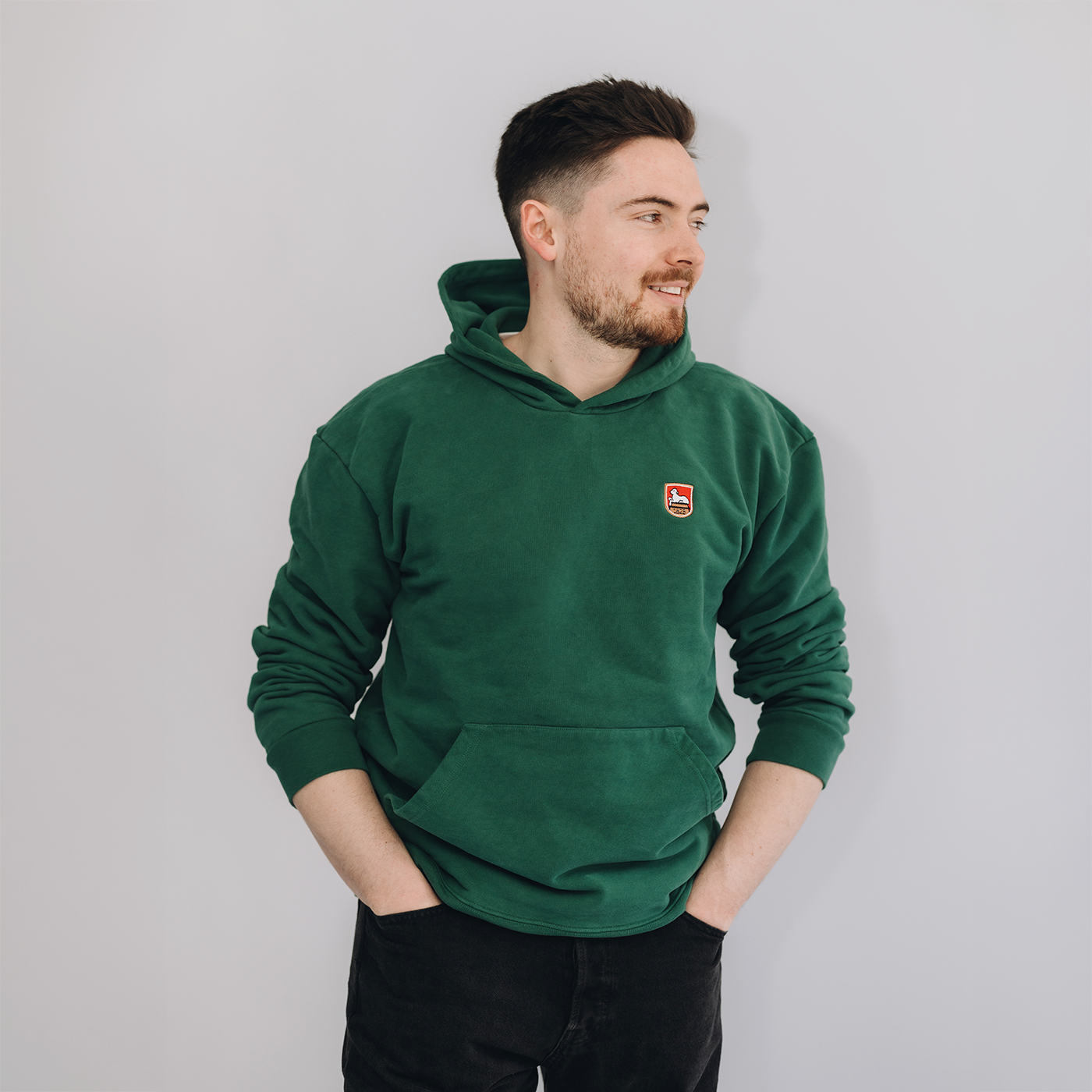Mann trägt grünes Kapuzen-Sweatshirt mit einem kleinen gestickten Lammsbräu-Wappen auf der Brust und schwarze Hose, Ansicht auf neutralem grauem Hintergrund. Er schaut nach links und lächelt, beide Hände in den Hosentaschen.