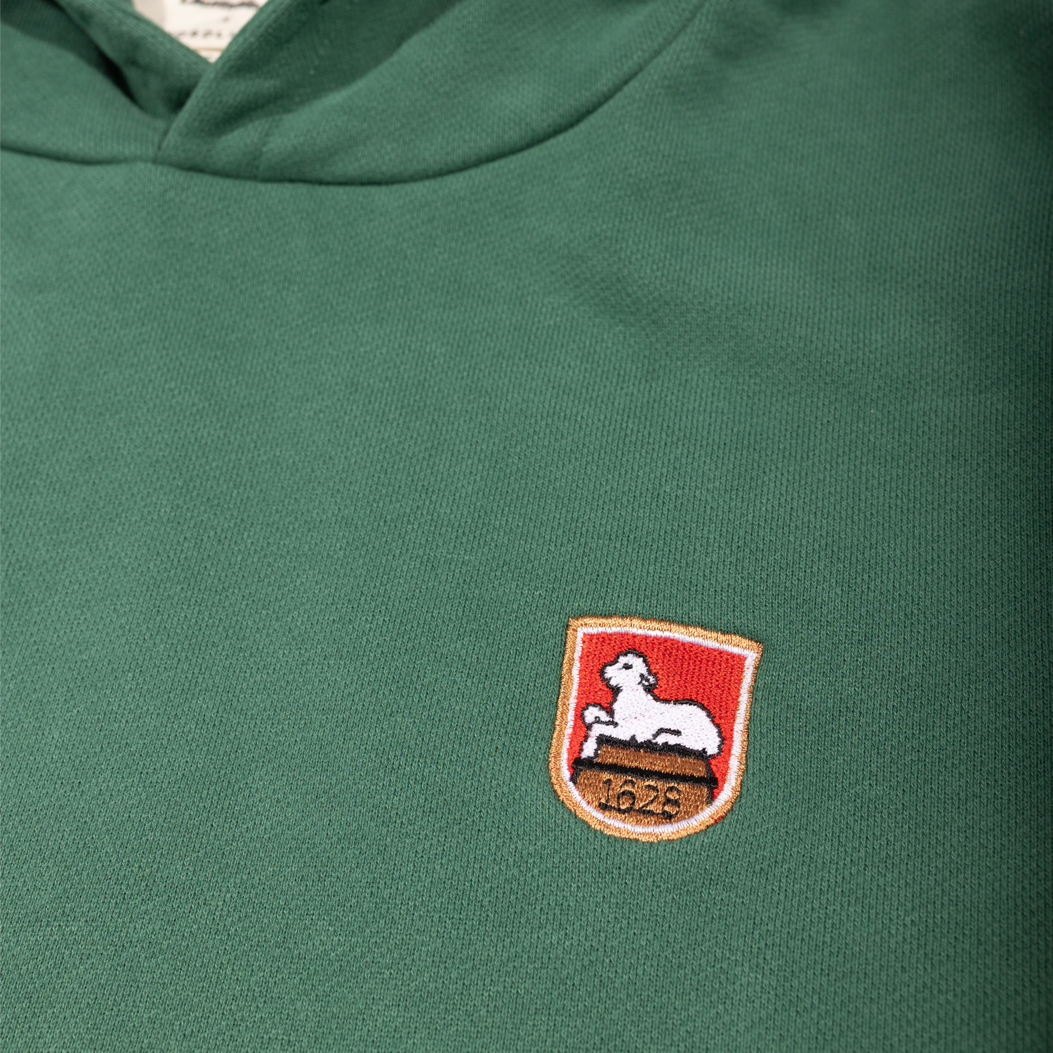 Detailaufnahme eines grünen Kapuzen-Sweatshirts mit gesticktem Wappen auf der linken Brustseite. Das Wappenschild zeigt ein weißes Lamm auf rotem Hintergrund, einen braunen Fassboden und die Jahreszahl 1628 mit goldenem Rand.