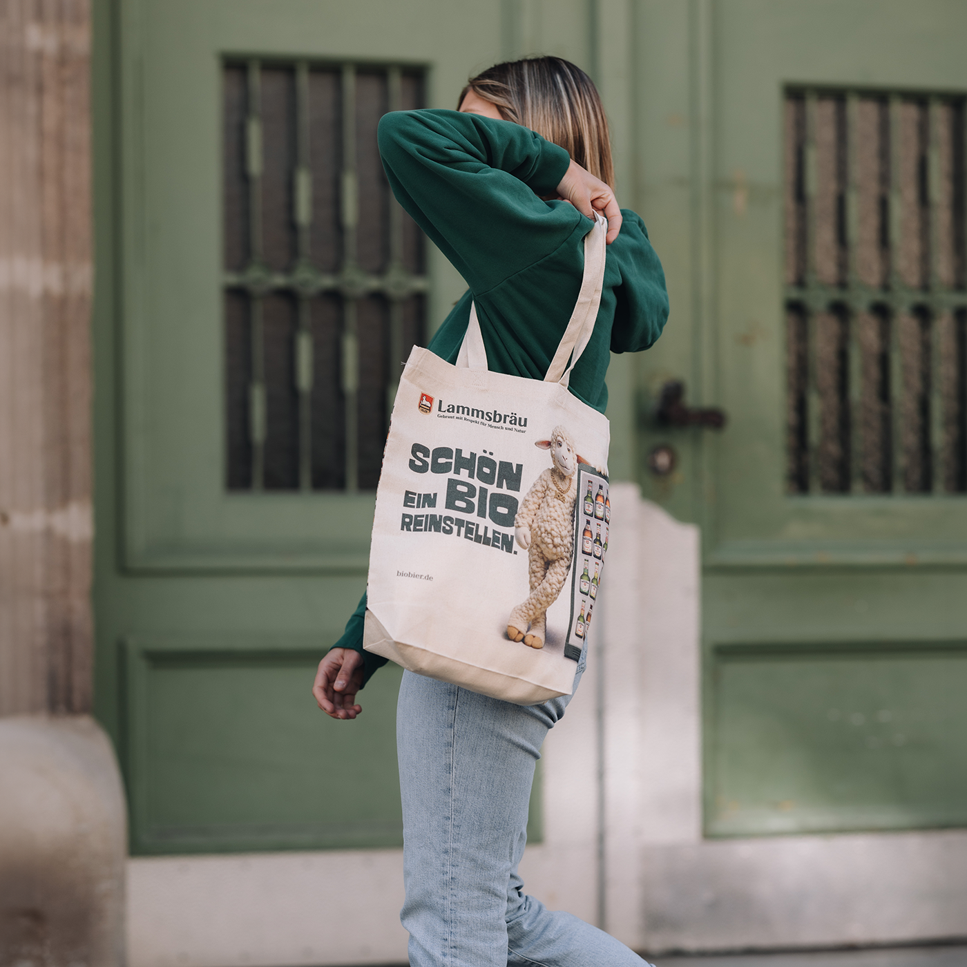 Frau trägt grünes Langarm-Sweatshirt und hellblaue Jeans vor einer grünen Gebäudefassade mit Fenstern. Sie trägt eine cremefarbene Baumwolltasche über der Schulter mit Lammsbräu-Logo, großem Lamm-Motiv und Text "SCHÖN EIN BIO REINSTELLEN" sowie einer Abbildung verschiedener Bierflaschensorten auf der rechten Seite. Ihre Hand hält die Tasche fest.