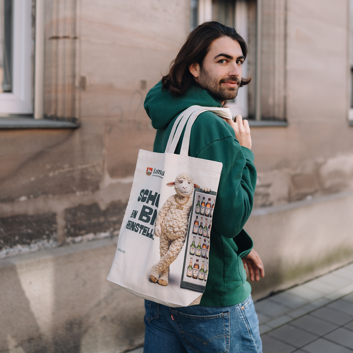 Mann mit dunklem Bart trägt grünes Kapuzen-Sweatshirt und blaue Jeans vor einer beigen Gebäudefassade. Er trägt eine cremefarbene Baumwolltasche über der Schulter mit Lammsbräu-Logo, Lamm-Motiv und Text "SCHÖN EIN BIO REINSTELLEN" sowie Bierflaschensorten-Abbildung. Er blickt über die Schulter zurück zur Kamera.