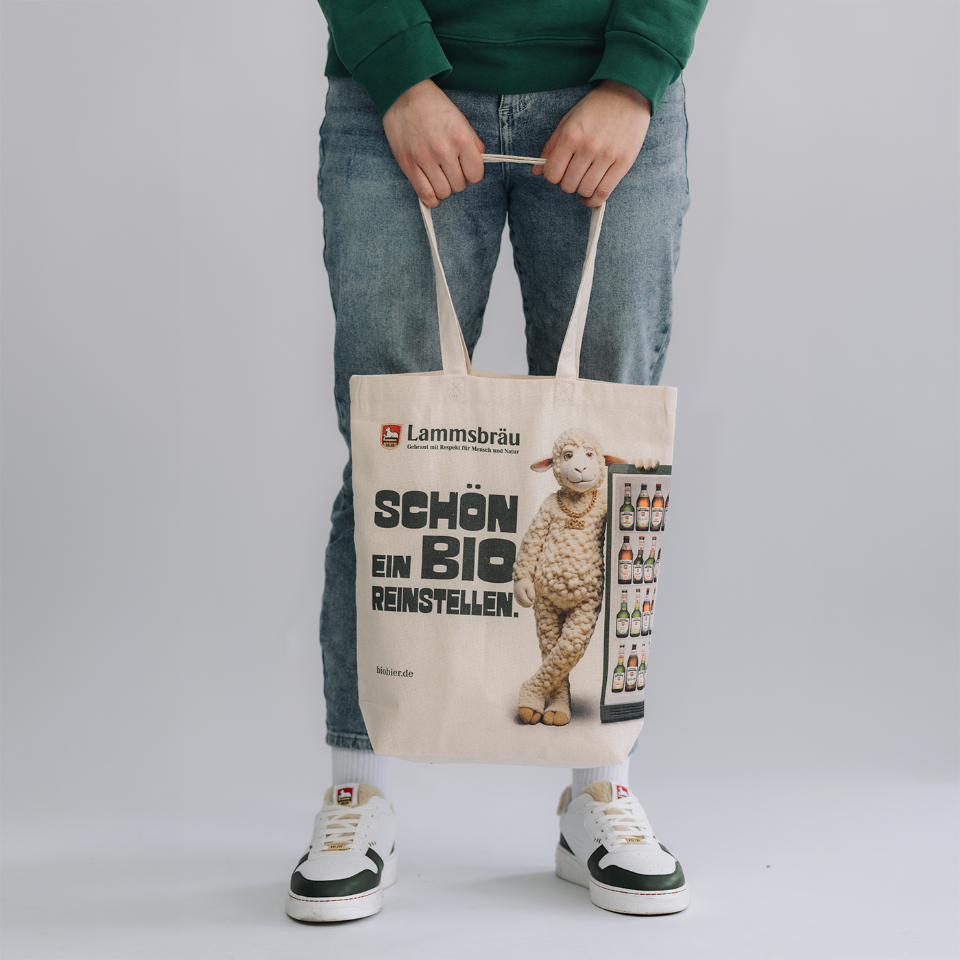 Person trägt grünes Sweatshirt und Jeans auf neutralem weißem Hintergrund. Die cremefarbene Baumwolltasche mit Lammsbräu-Logo, großflächigem Lamm-Motiv in Vorderansicht und Text "SCHÖN EIN BIO REINSTELLEN" wird mit beiden Händen von oben gehalten. Darunter sind weiße Sneaker mit grünen Akzenten sichtbar.
