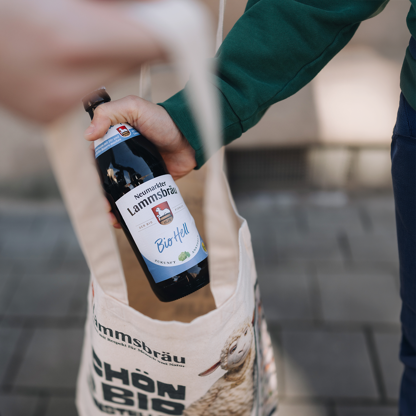Person in grünem Sweatshirt und cremefarbener Baumwolltasche hält eine braune Lammsbräu-Bierflasche mit blauem Etikett "Bio Hell" in der Hand. Das Etikett zeigt das Lammsbräu-Wappen oben, darunter "Neumarkter Lammsbräu" und "BioHell" in blauer Schrift. Die Tasche trägt das Lammsbräu-Logo und Text "SCHÖN EIN BIO REINSTELLEN". Im Hintergrund verschwommene Gebäudefassade.