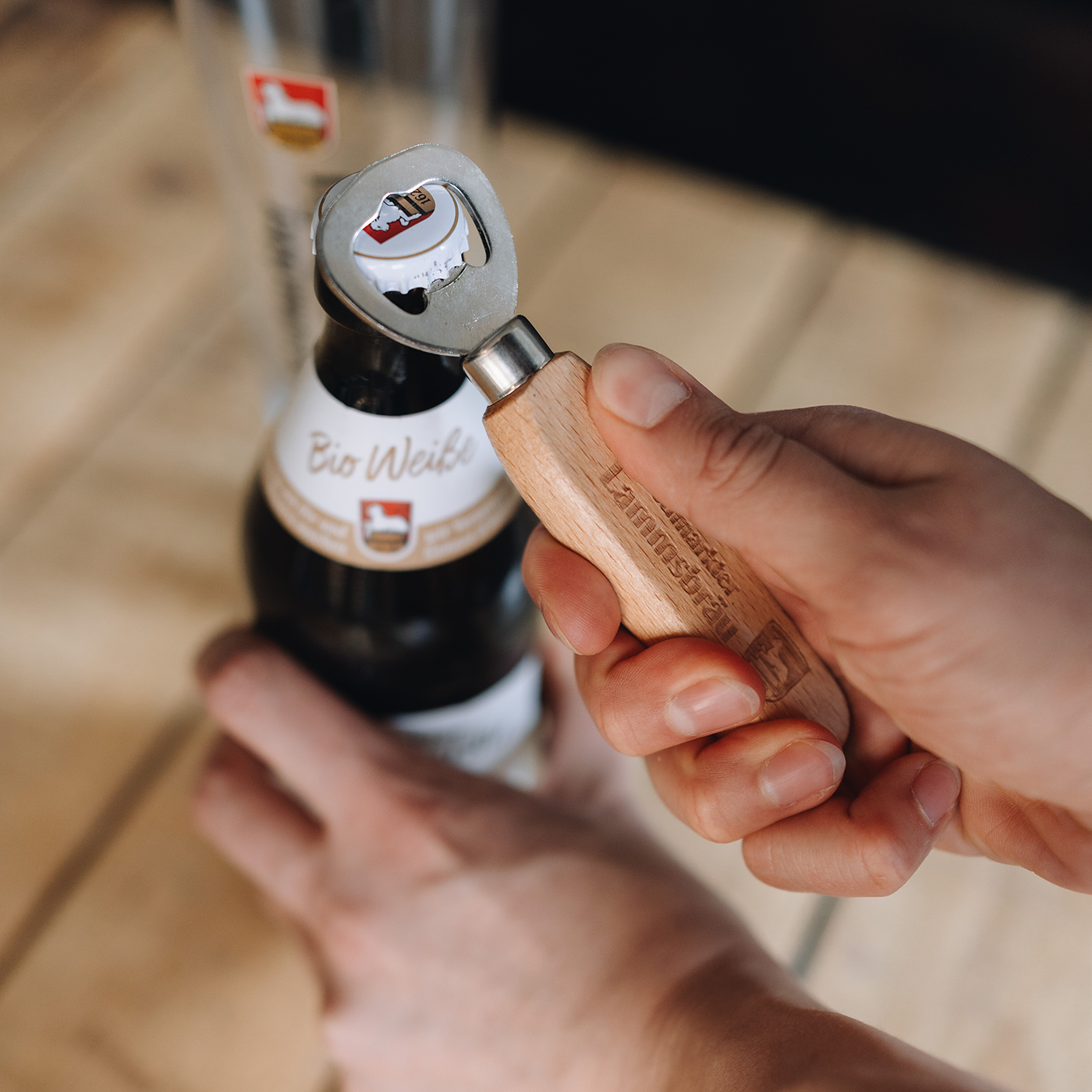 Eine Hand hält einen Bieröffner mit Griff aus hellem Holz und rundem Metallkopf. Im Hintergrund unscharf gestellt eine dunkle Bierflasche mit weißem Etikett und rotem Wappenlabel. Der Öffner wird in Nahaufnahme von vorne präsentiert auf Holzuntergrund.