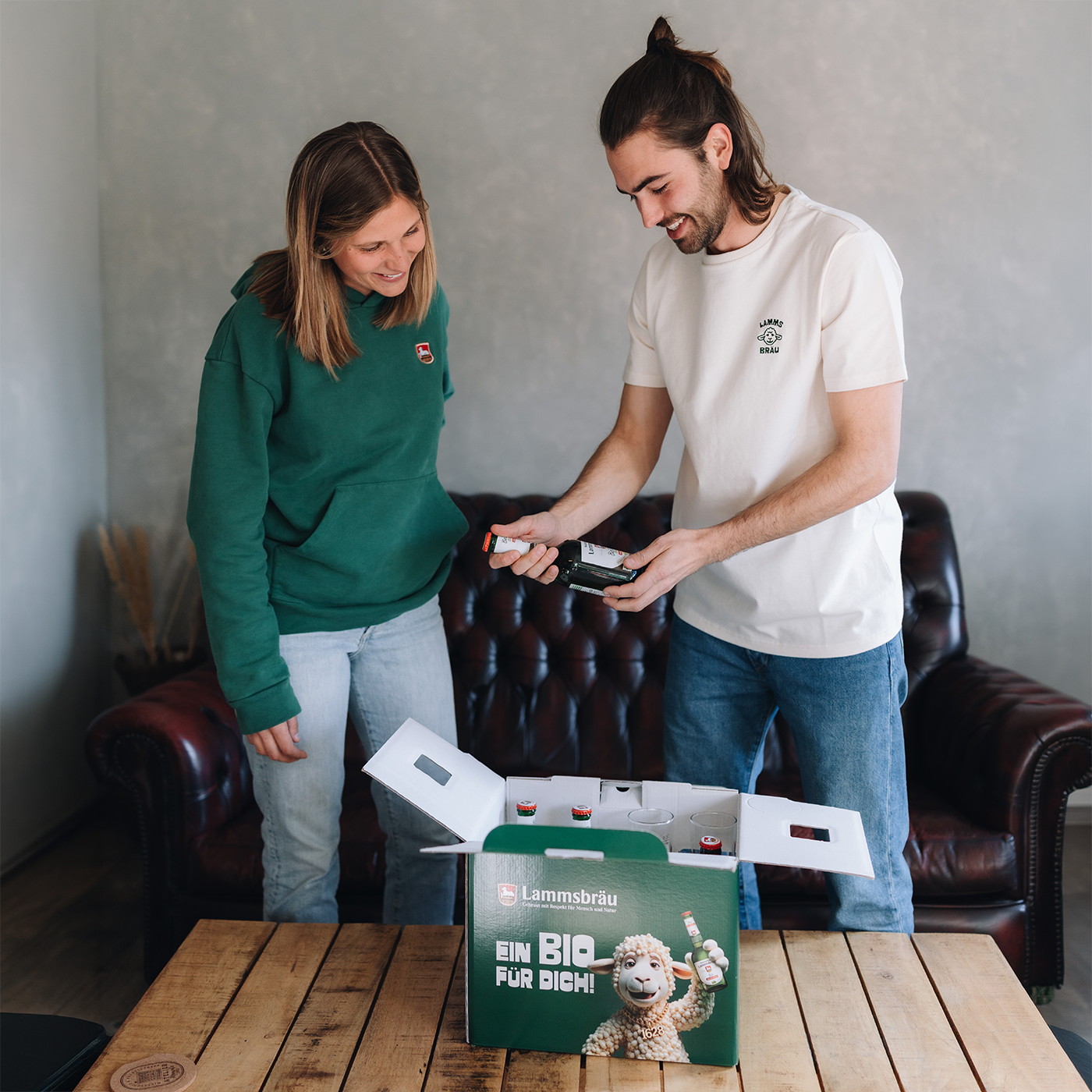 Frau in dunkelgrünem Sweatshirt mit kleinem Lammsbräu-Logo und hellblauer Jeans und Mann in weißem T-Shirt mit dunkelgrünem Lammsbräu-Motiv und dunkelblauer Jeans stehen vor einem dunklen Ledersofa. Sie betrachten zusammen eine große grüne Lammsbräu-Geschenkbox mit weißem Text "EIN BIO FÜR DICH!" und einem stilisierten weißen Lamm-Motiv.