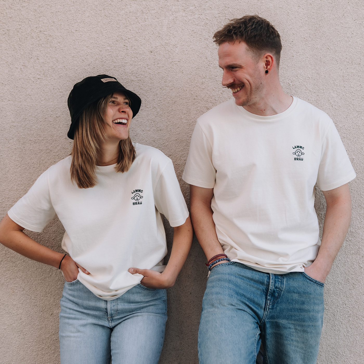 Eine Frau und ein Mann tragen jeweils ein weißes T-Shirt mit einem kleinen dunkelgrünen Lammsbräu-Motiv auf der Brust. Beide kombinieren das Shirt mit hellblauer Jeans. Die Frau trägt zusätzlich einen dunkelgrünen Bucket Hat. Sie stehen nebeneinander vor einer beigen Wand und blicken sich lächelnd an.