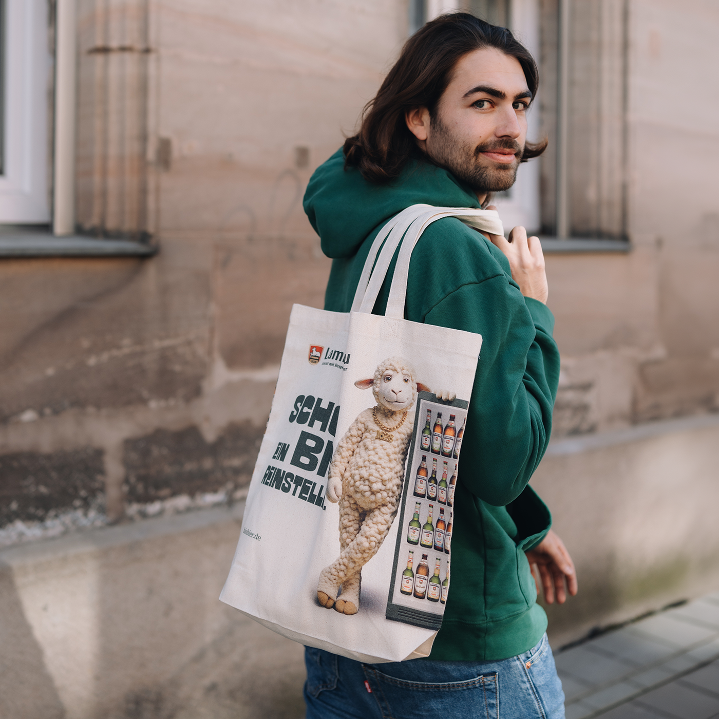 Ein Mann trägt über der Schulter eine cremefarbene Baumwoll-Tragetasche mit langen Henkeln. Das Motiv zeigt ein beiges Lamm in Frontalansicht, daneben Text und eine Rasterdarstellung von Bierflaschen. Der Mann trägt einen dunkelgrünen Kapuzenpullover und blaue Jeans und blickt über die Schulter zurück. Die Tasche wird in seitlicher Trageposition vor einer Backsteinwand fotografiert.