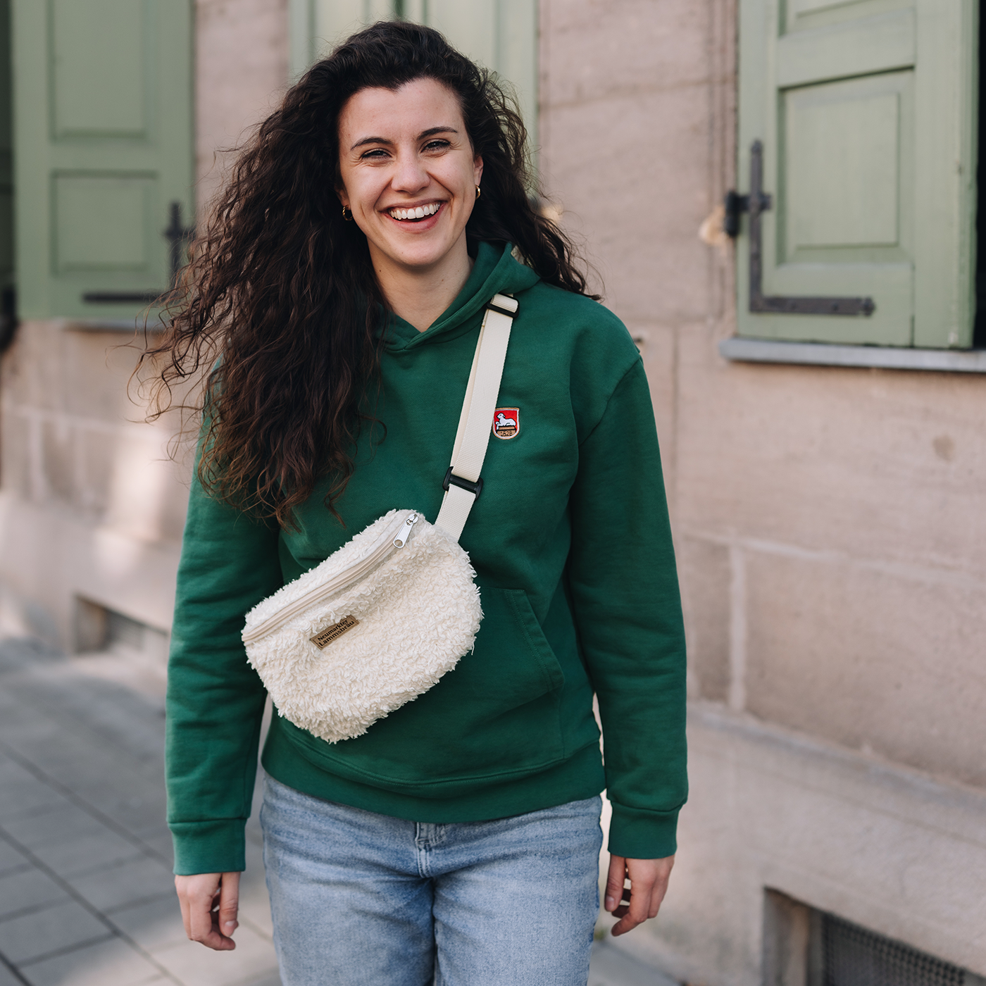 Frau trägt grünes Kapuzen-Sweatshirt mit kleinem gestickten Lammsbräu-Wappen auf der Brust und hellblauer Jeans. Sie trägt die cremefarbene Umhängetasche mit weißem Gurt über der Schulter vor einer historischen Gebäudefassade mit grünen Türen.