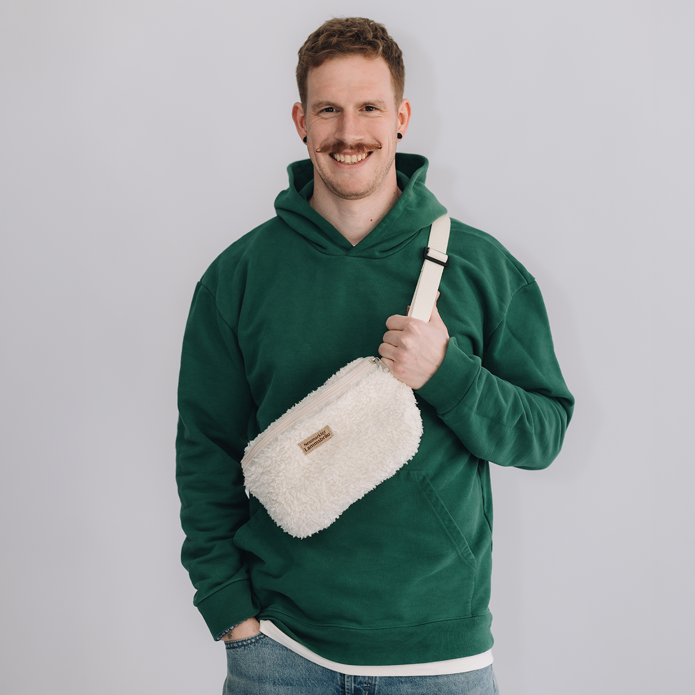 Mann trägt grünes Kapuzen-Sweatshirt mit cremefarbene Umhängetasche und hellblauer Jeans auf neutralem grauem Hintergrund. Er hält die cremefarbene Umhängetasche mit weißem Gurt und braunem Leder-Label in der Hand, lächelt in die Kamera.