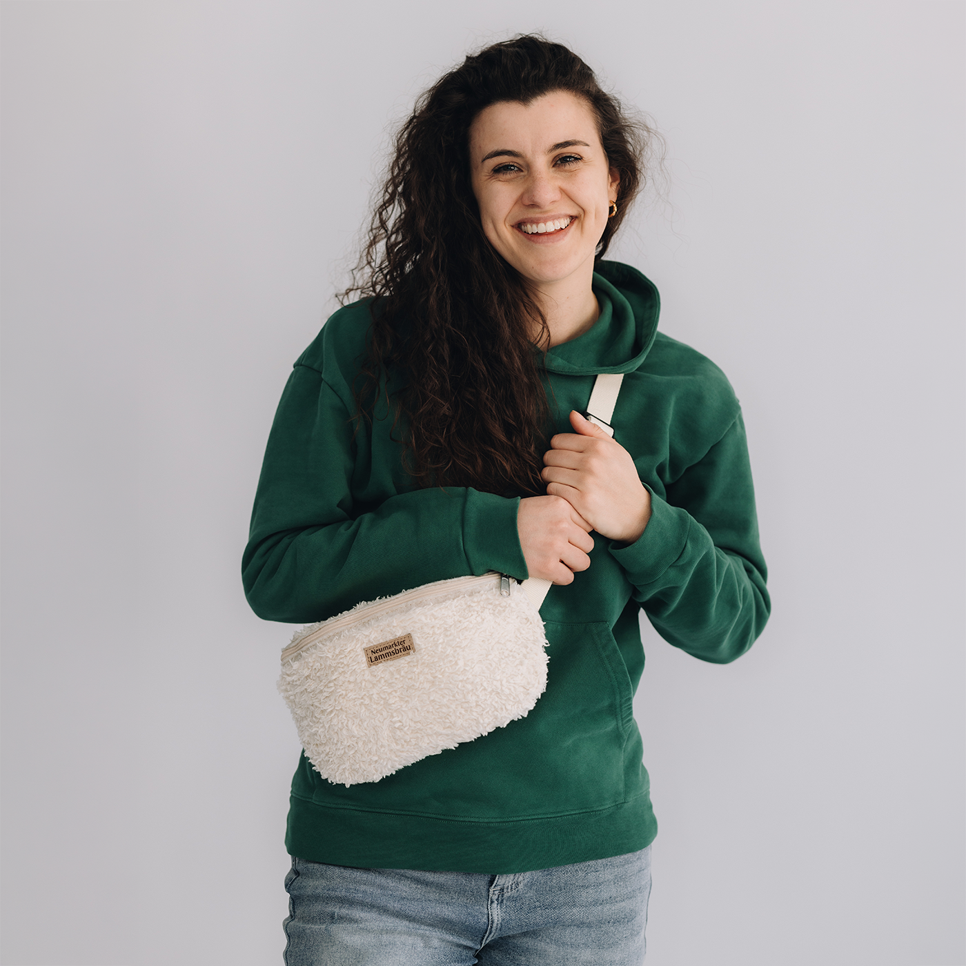 Frau trägt grünes Kapuzen-Sweatshirt mit cremefarbener Umhängetasche und Jeans auf neutralem grauem Hintergrund. Sie hält die cremefarbene Umhängetasche mit weißem Gurt und braunem Leder-Label vor sich, lächelt in die Kamera.