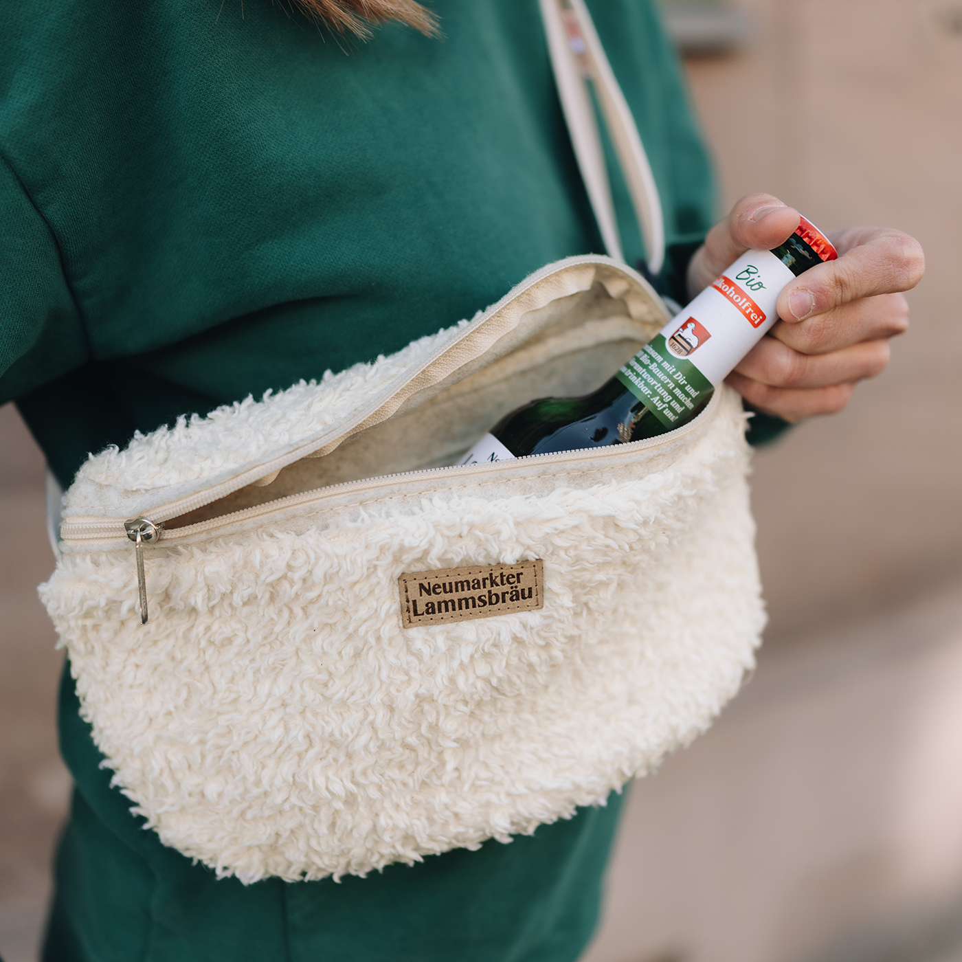 Frau trägt grünes Langarm-Sweatshirt und trägt eine offene cremefarbene-Umhängetasche. Aus der geöffneten Tasche ragt eine grüne Lammsbräu-Bierflasche mit rotem Kronkorken heraus. Das beige Leder-Label mit "Neumarkter Lammsbräu"-Schriftzug ist sichtbar. Im Hintergrund verschwommene urbane Kulisse.