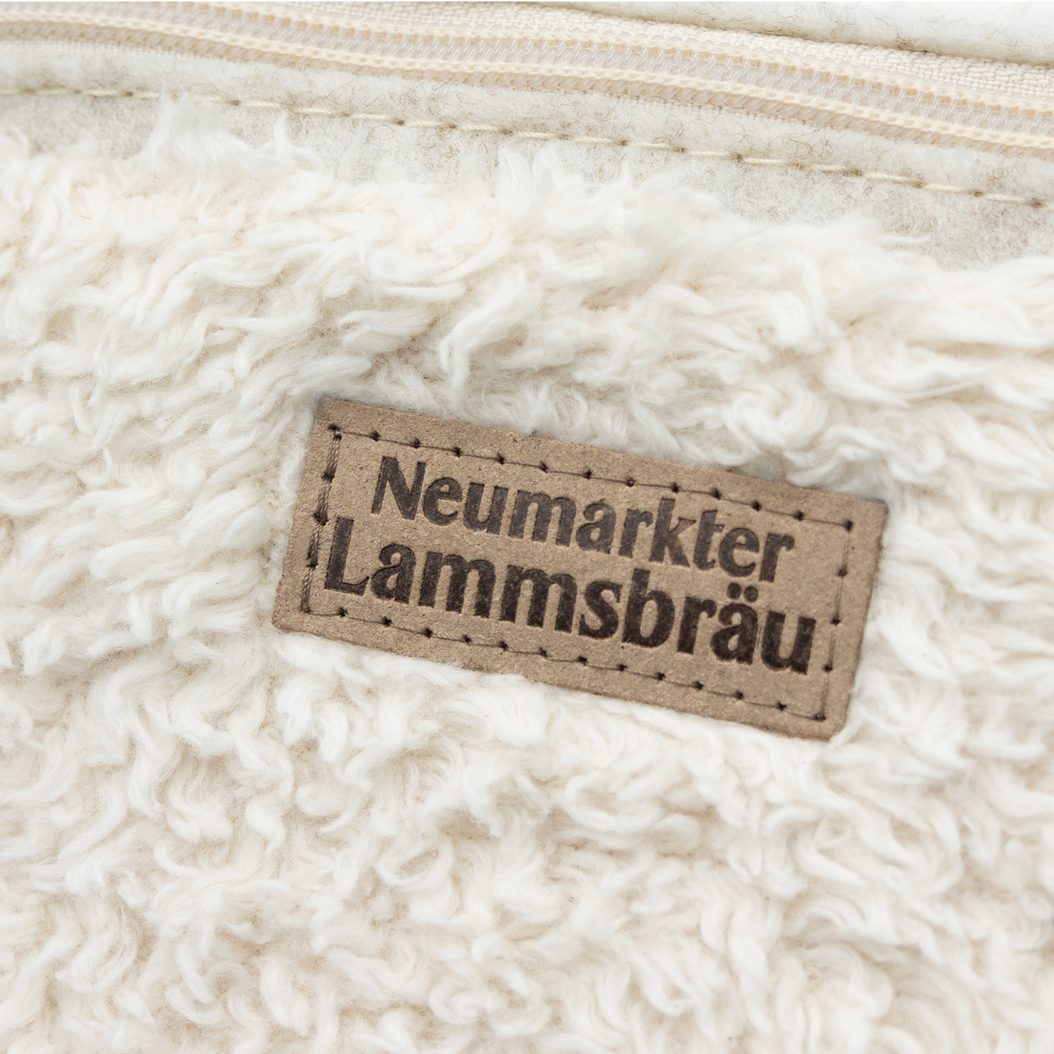 Detailaufnahme einer cremefarbenen Umhängetasche mit strukturierter Oberfläche. Ein rechteckiges beiges Leder-Label mit schwarzer Schrift "Neumarkter Lammsbräu" ist mittig auf der Tasche angebracht und mit dekorativen Stichlinien verziert.