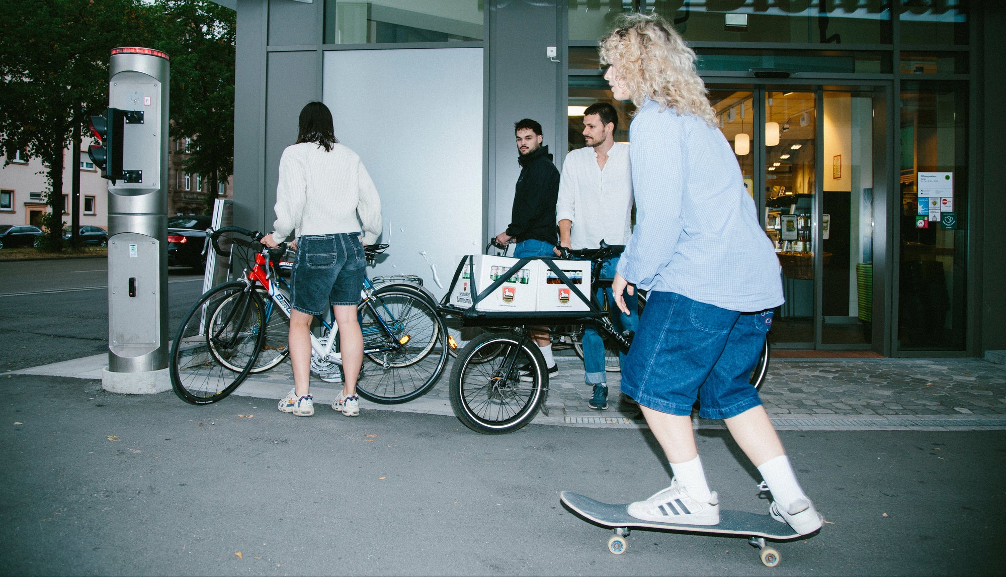 Vier Personen befinden sich mit Fahrrädern und einem Lastenrad vor dem Eingang eines Gebäudes an einer Straße. Im Vordergrund fährt eine Person mit lockigem blondem Haar auf einem Skateboard vorbei; links steht eine Ampelanlage am Straßenrand.