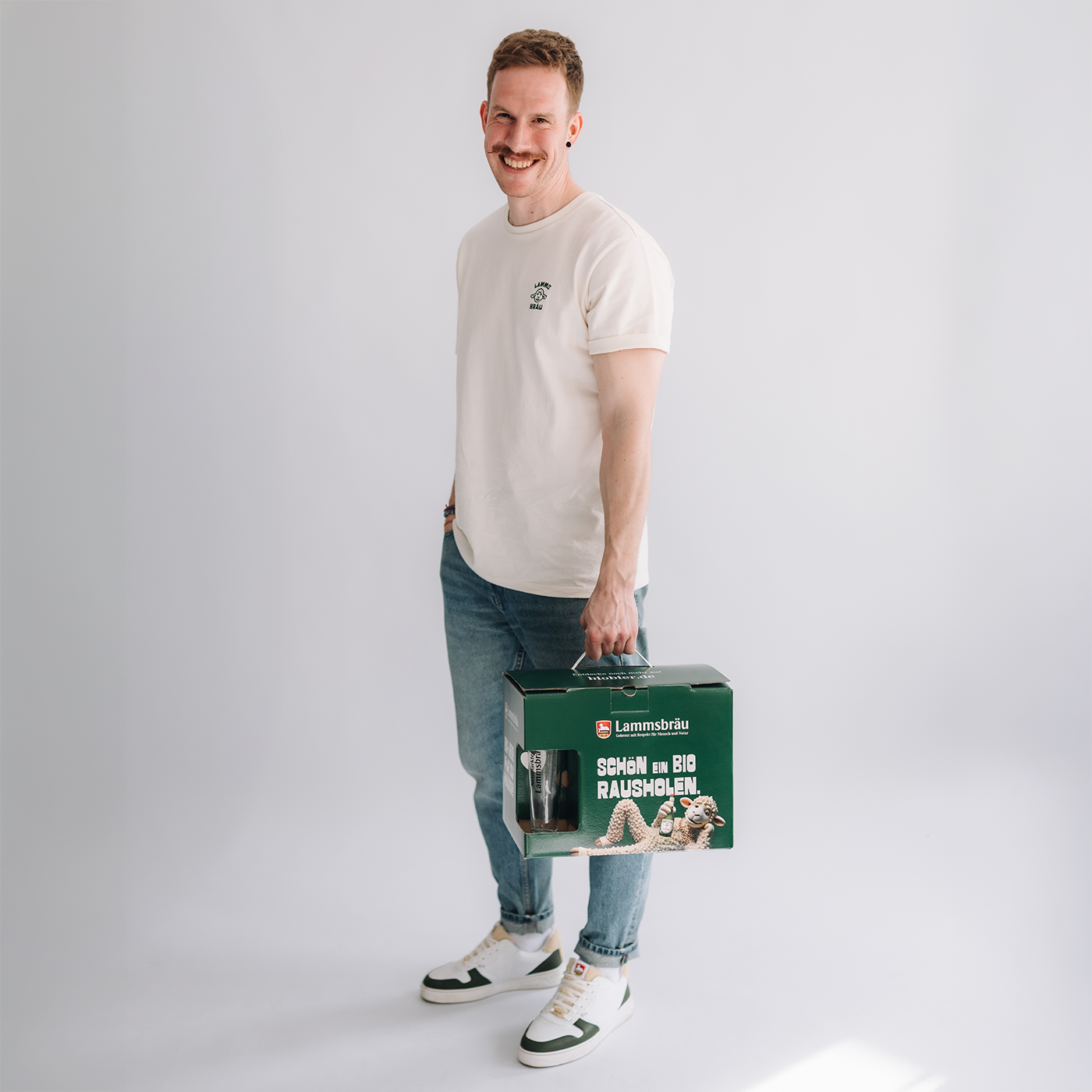 Mann mit braunem Haar trägt cremefarbenes Kurzarm-T-Shirt mit kleinem Lammsbräu-Motiv auf der Brust und blaue Jeans vor neutralem grauem Hintergrund. Er hält eine grüne Lammsbräu-Geschenkbox mit weißem Text "SCHÖN EIN BIO RAUSHOLEN" und Lamm-Motiv in der rechten Hand, lächelt in die Kamera.
