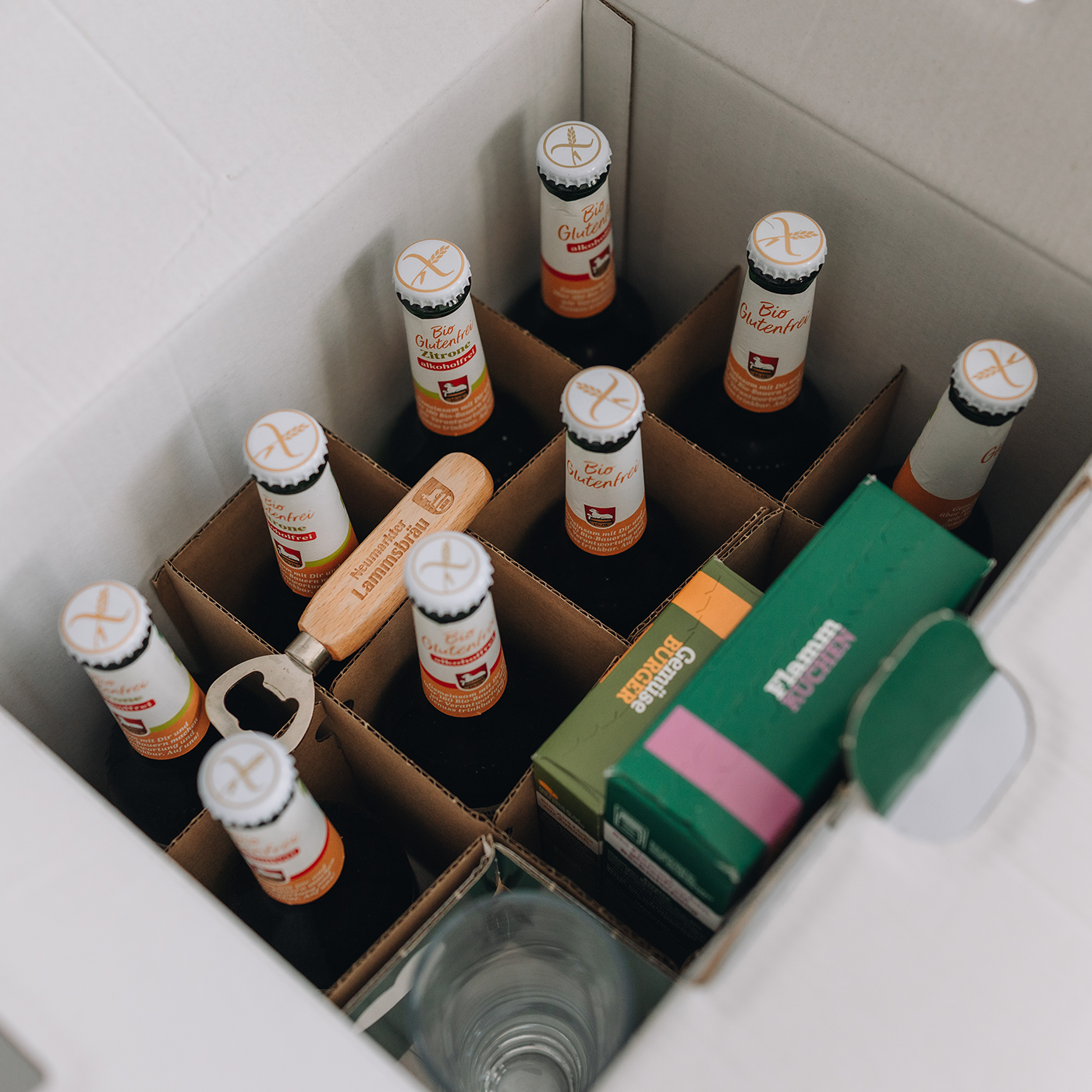 Offene weiße Paketbox von oben mit Trennwänden, gefüllt mit neun Lammsbräu-Bierflaschen mit weißem Kronkorken. In der Mitte liegt ein Holz-Flaschenöffner mit eingraviertem Lammsbräu-Logo. Rechts liegen grüne 
Verpackungsboxen, unten sichtbar ist ein Glas.