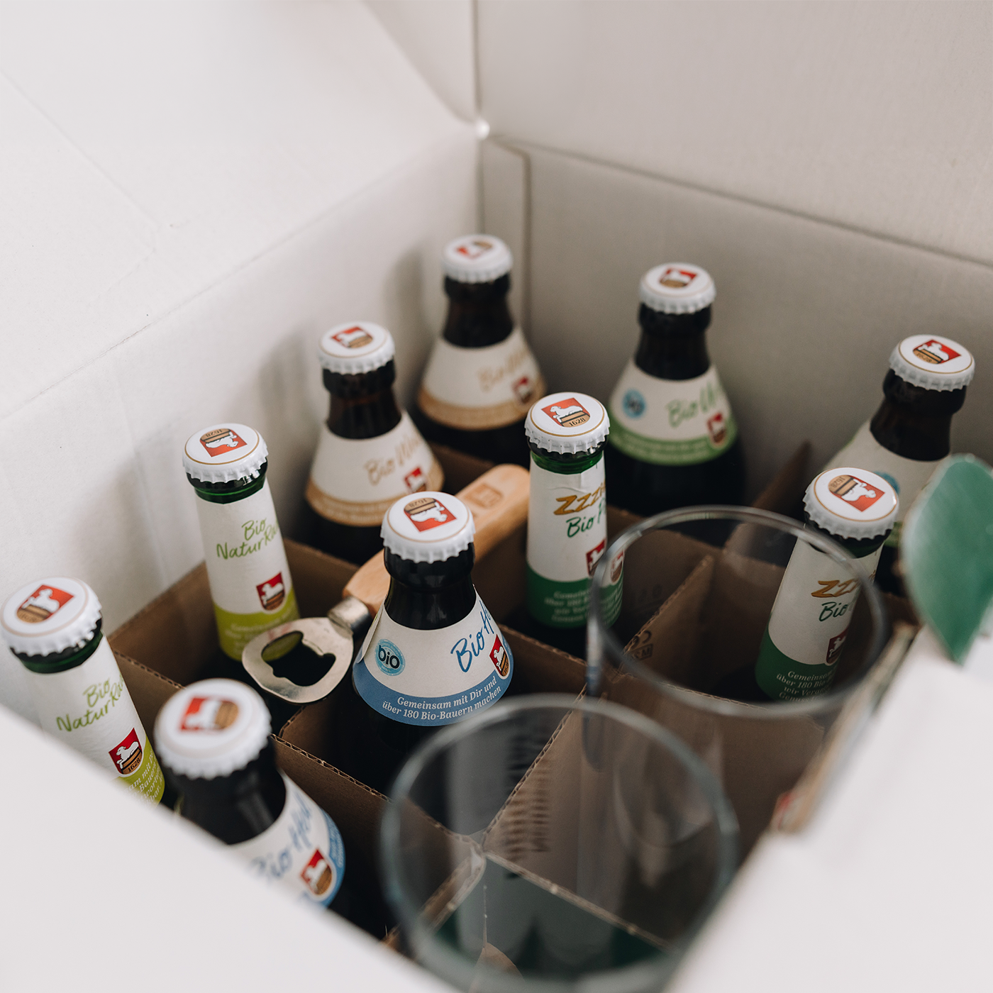 Offene weiße Paketbox von oben mit braunen Trennwänden in perspektivischer Nahaufnahme. In der Box sind 10 Bierflaschen. In der Mitte der Box ist ein Holz-Flaschenöffner.  Rechts zwei transparente Gläser mit Lammsbräu-Schriftzug.