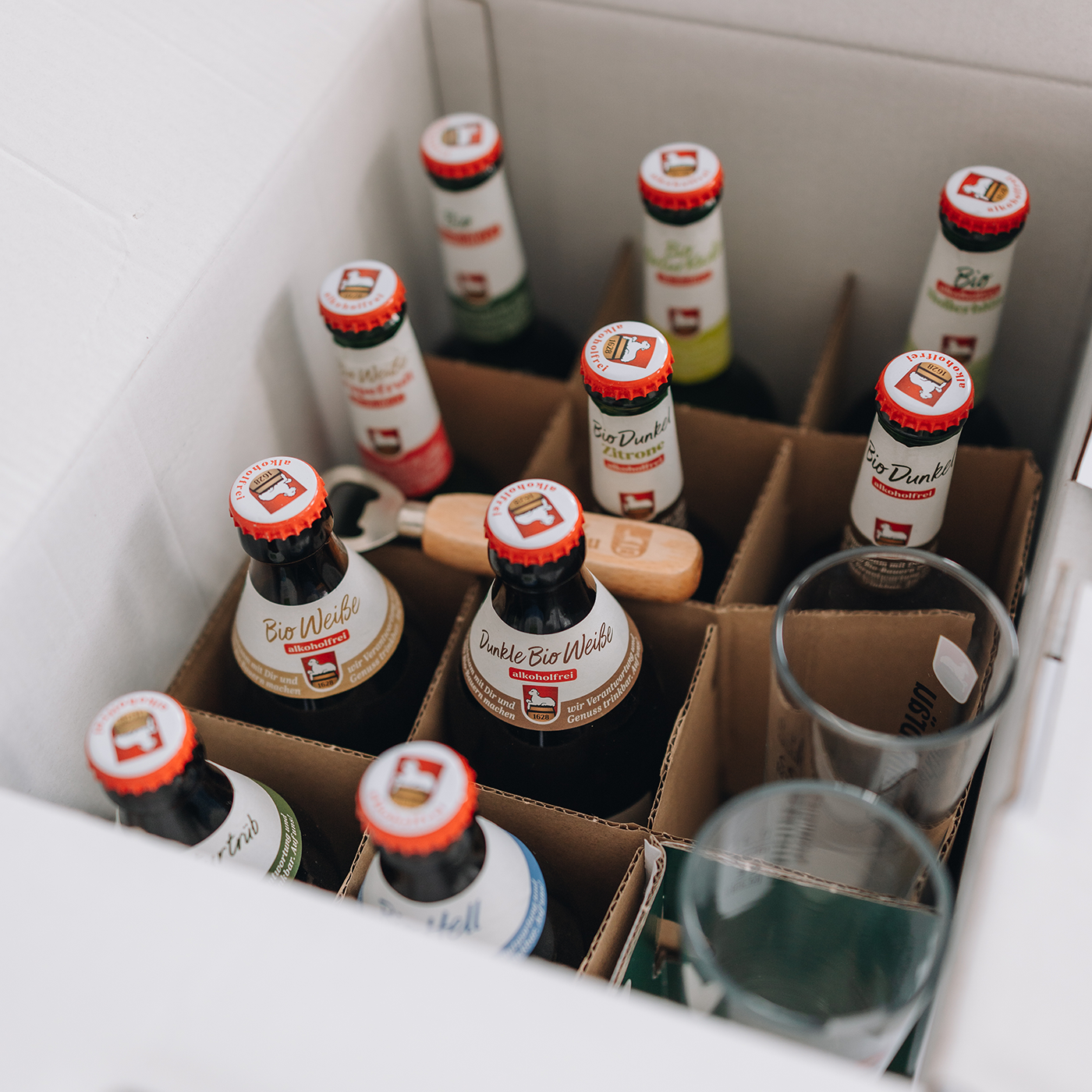 Offene weiße Paketbox von oben mit braunen Trennwänden, gefüllt mit verschiedenen Lammsbräu-Bierflaschen mit rot-weißen Kronkorken. In der Ecke rechts zwei transparente Gläser mit Lammsbräu-Druck. In der Bildmitte liegt ein beiger Holz-Flaschenöffner mit eingraviertem Lammsbräu-Logo.