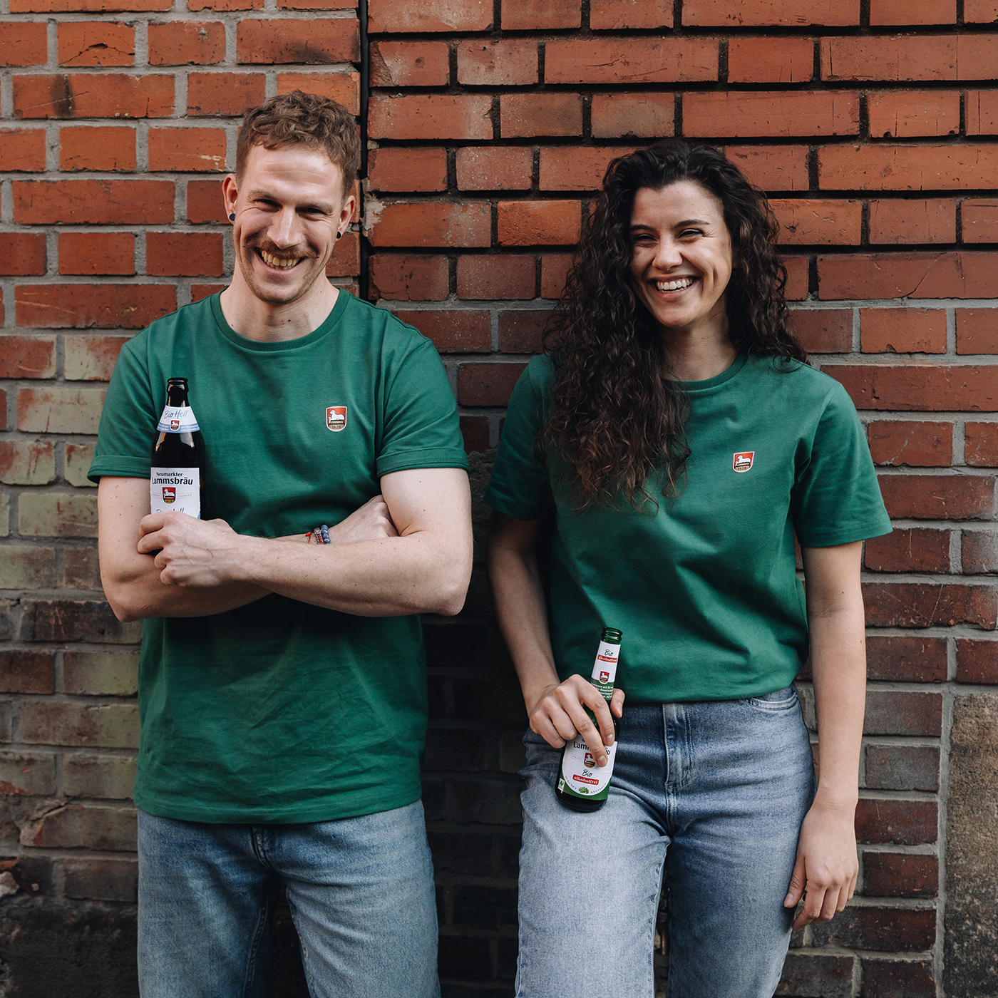Mann und Frau stehen vor einer roten Backsteinwand, beide tragen grüne T-Shirts mit kleinem Lammsbräu-Wappen auf der Brust und hellblaue Jeans. Beide halten jeweils eine Lammsbräu-Bierflasche in der Hand und lachen in die Kamera.