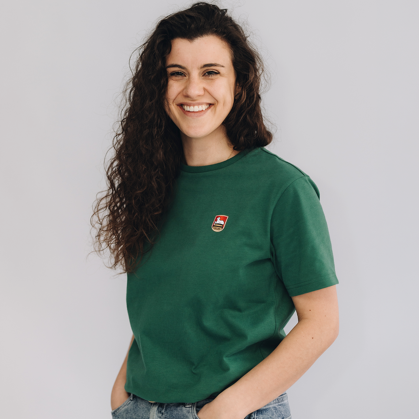 Frau trägt grünes Kurzarm-T-Shirt mit kleinem Lammsbräu-Wappen auf der Brust. Die Frau steht vor einem neutralen grauen Hintergrund.