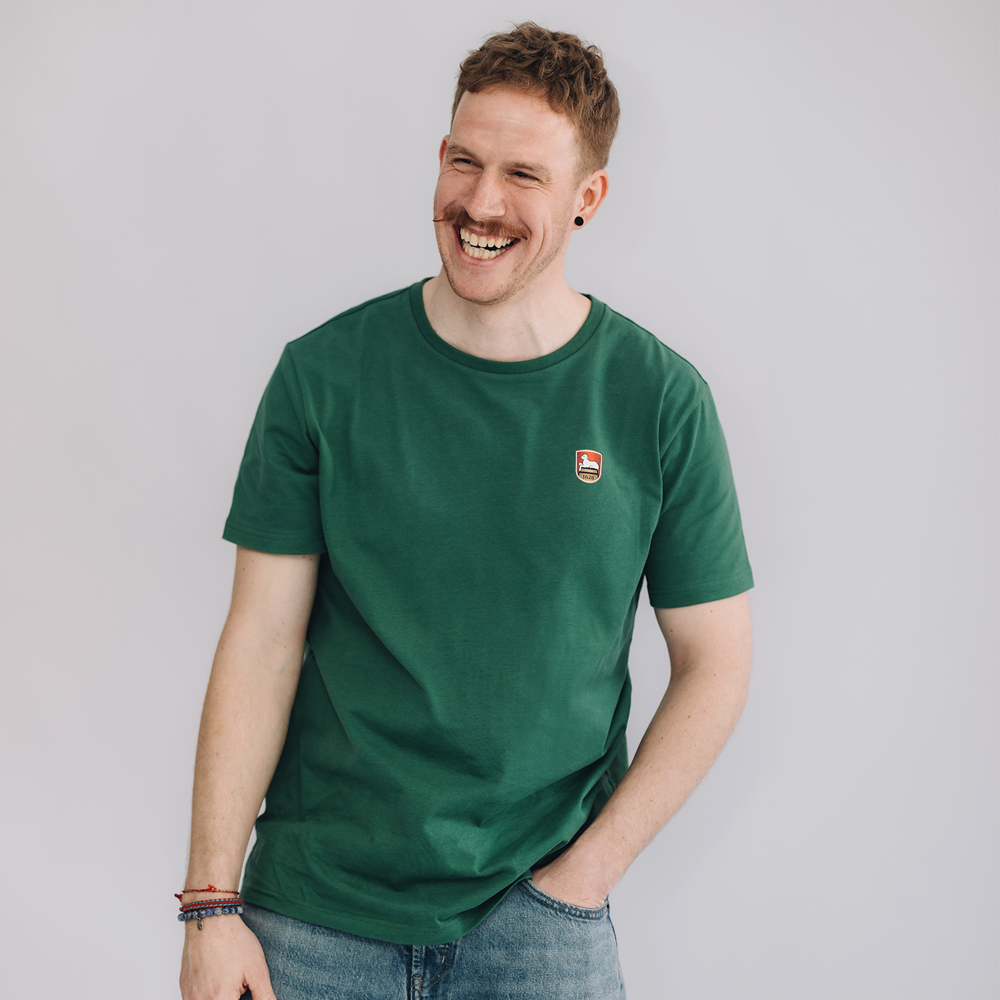 Mann trägt grünes Kurzarm-T-Shirt mit kleinem Lammsbräu-Wappen auf der Brust und hellblaue Jeans. Der Mann steht vor einem neutralen grauen Hintergrund. 
