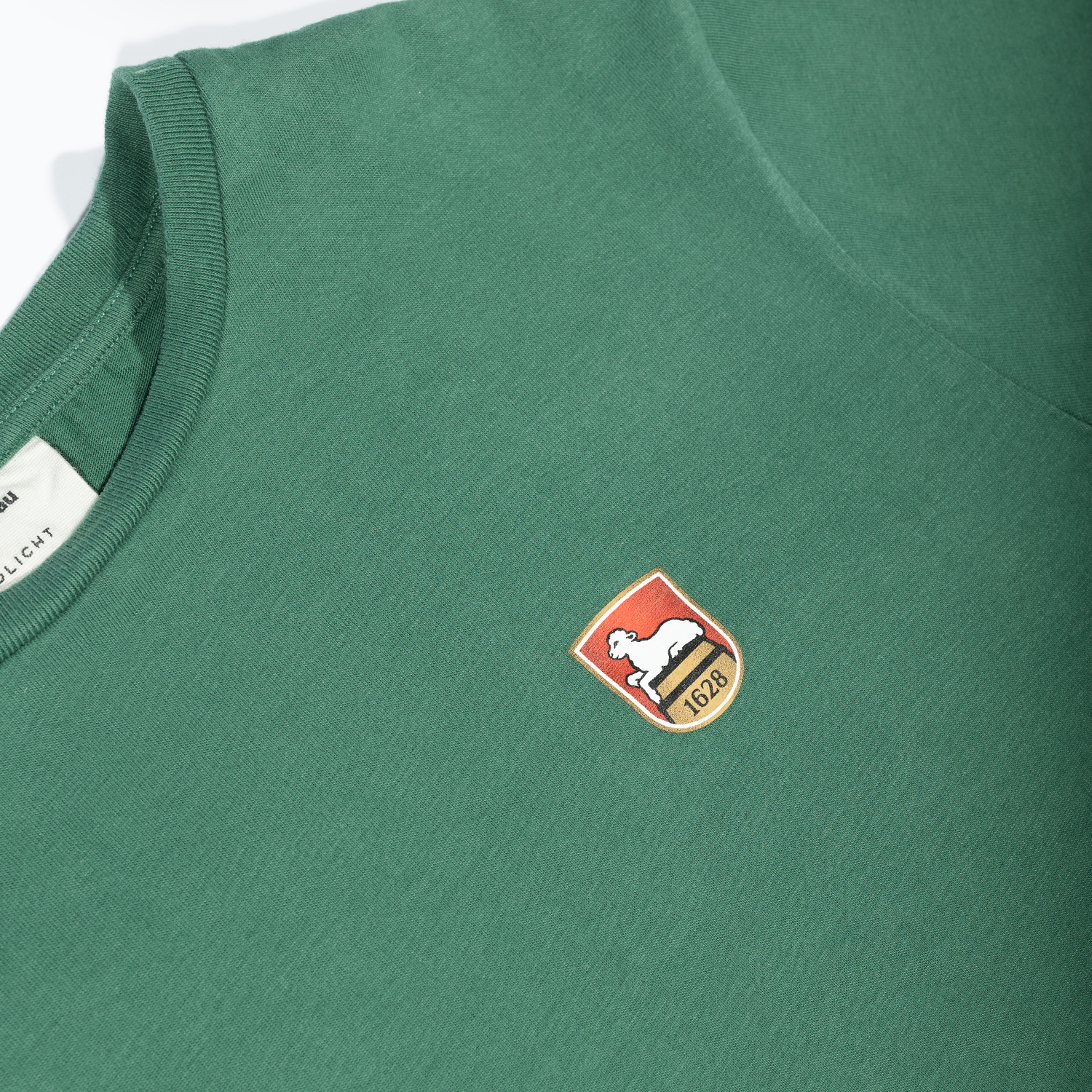 Detailaufnahme eines grünen T-Shirts mit geripptem Rundhalsausschnitt. Auf der Brustpartie befindet sich das gedruckte Lammsbräu-Wappen, welches ein weißes Lamm auf rotem Grund sowie die Jahreszahl 1628 zeigt.