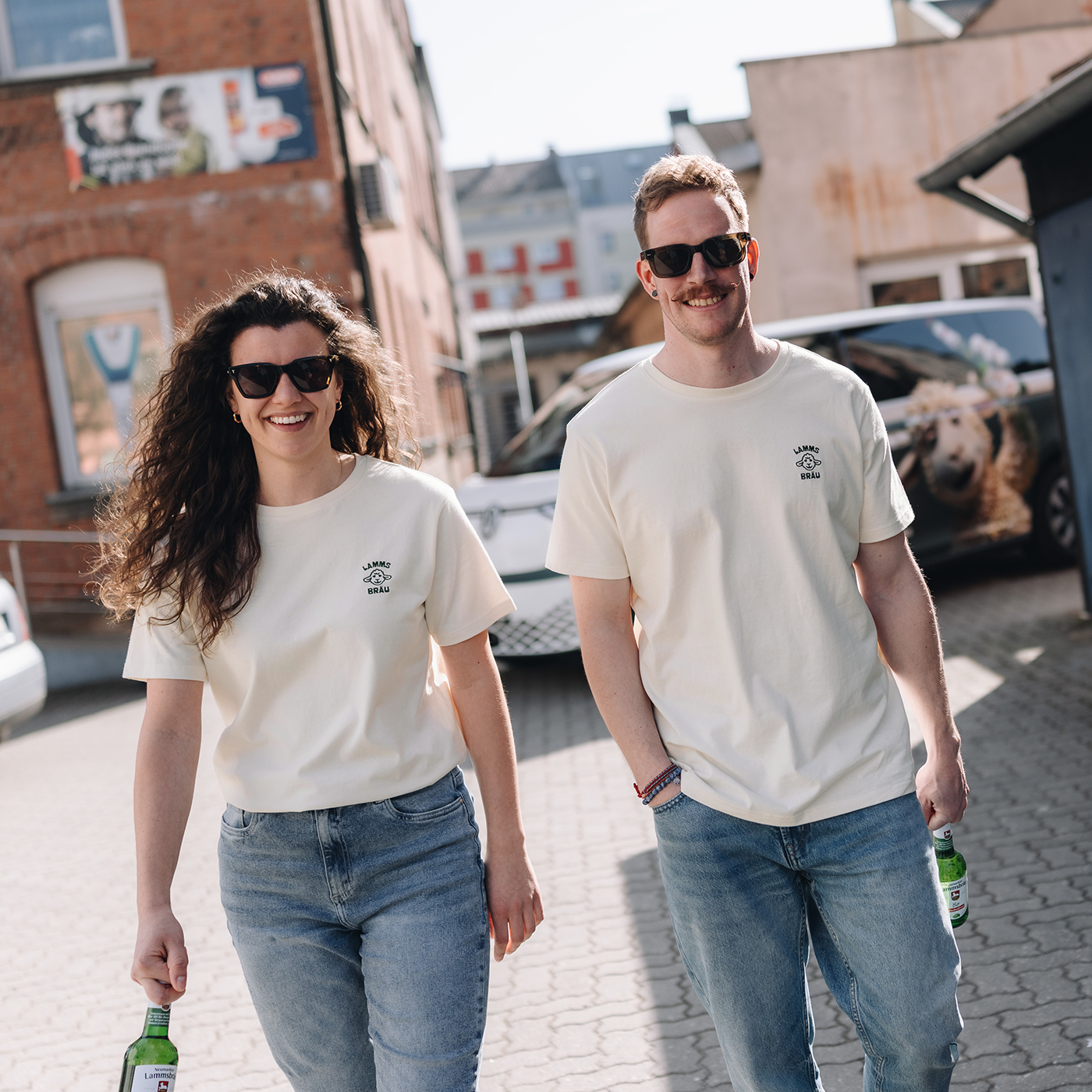 Frau und Mann gehen nebeneinander auf einem Kopfsteinpflaster-Platz vor roten Backsteingebäuden. Beide tragen weiße T-Shirts mit einem dunkelgrünen gestickten Lammsbräu-Motiv auf der Brust und hellblaue Jeans. Beide tragen Sonnenbrillen und halten jeweils eine Bierflasche in der Hand. 
