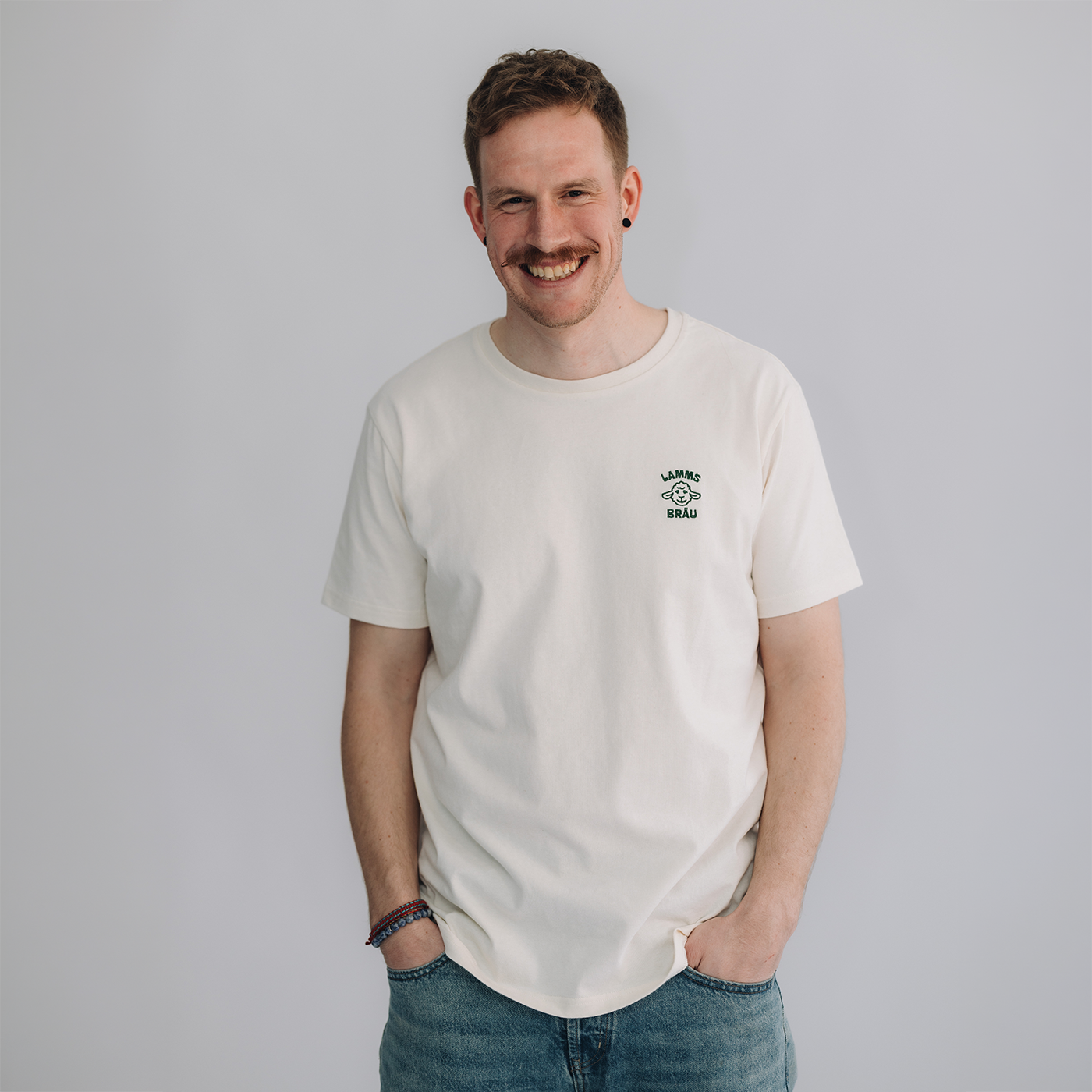 Mann trägt weißes Kurzarm-T-Shirt mit kleinem dunkelgrün gestickten Lammsbräu-Motiv und Schriftzug auf der Brust, dazu hellblaue Jeans. Er steht frontal vor neutralem grauem Hintergrund und lächelt in die Kamera.