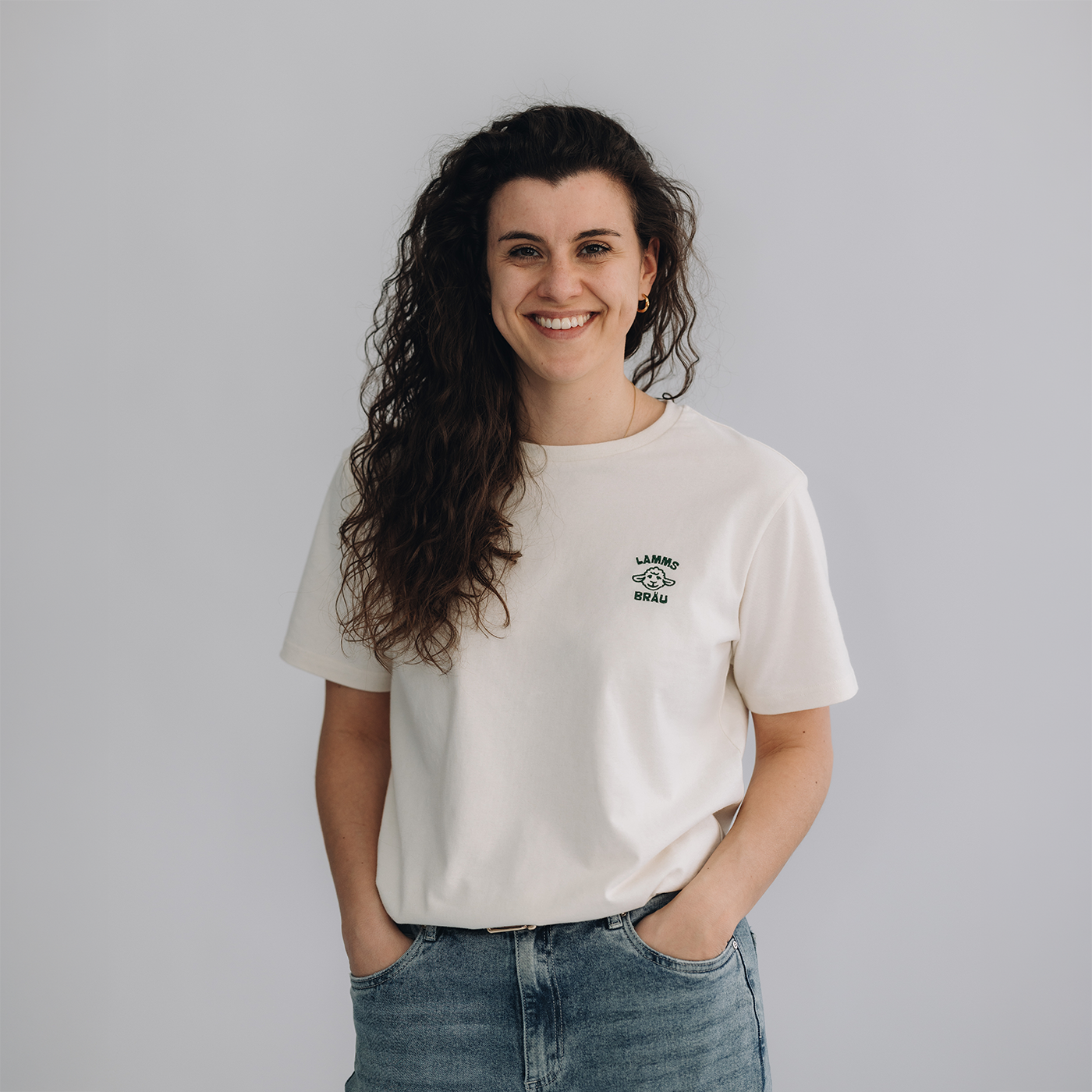 Frau trägt weißes Kurzarm-T-Shirt mit einem kleinen dunkelgrün gestickten Lammsbräu-Motiv und Schriftzug auf der Brust, dazu hellblaue Jeans. Sie steht frontal vor neutralem grauem Hintergrund und lächelt in die Kamera.