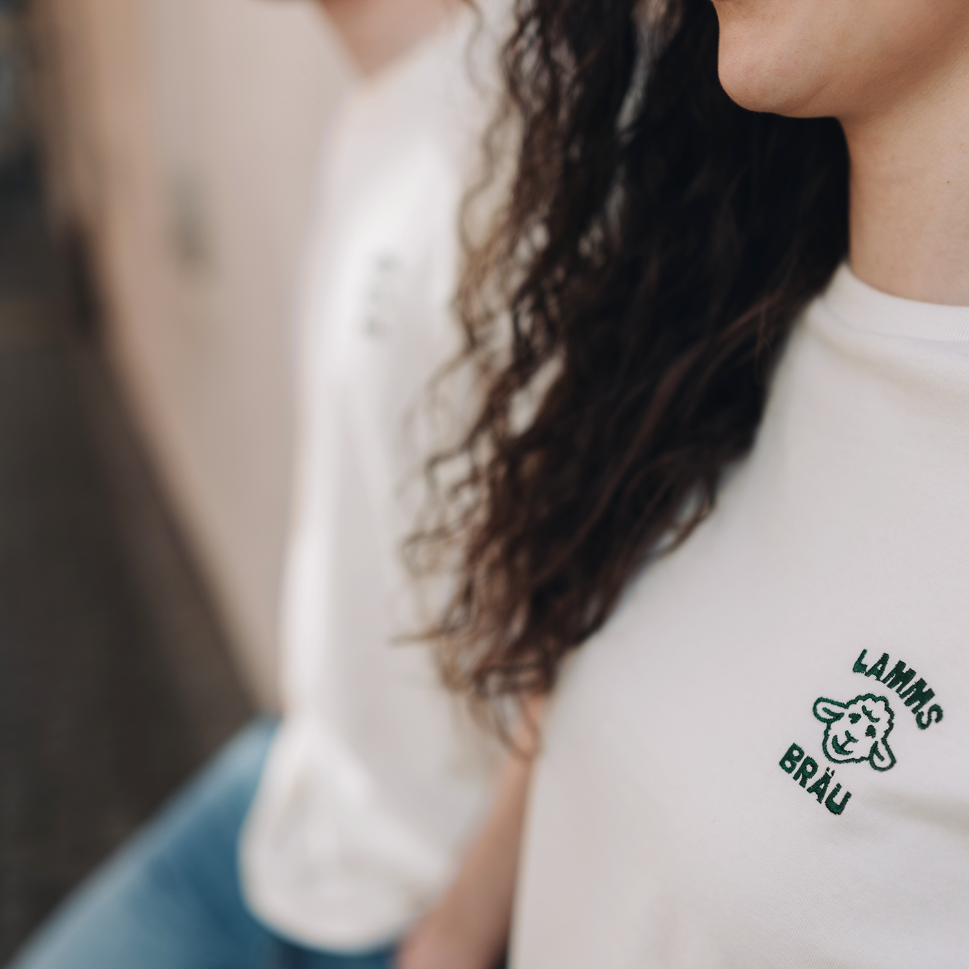 Nahaufnahme einer Person mit lockigem braunem Haar, die ein weißes T-Shirt trägt. Auf der rechten Brustseite ist ein grünes Lamm-Motiv und Schriftzug gestickt. Im Hintergrund ist verschwommen eine weitere Person in hellem T-Shirt sichtbar.